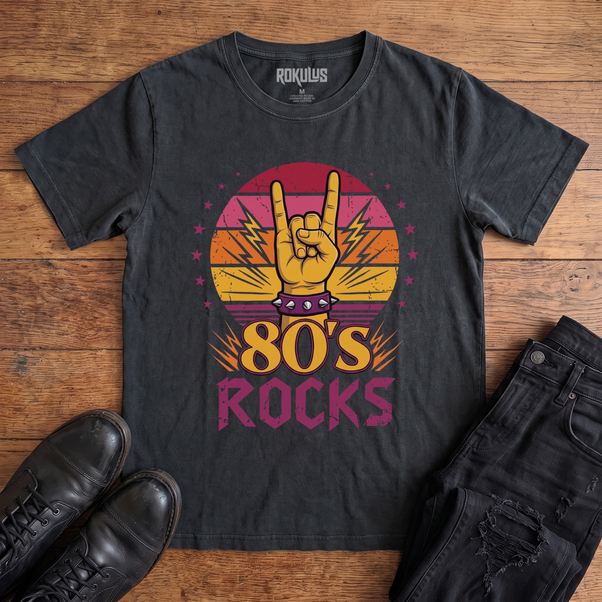 Washed black Rokulus 80's Rocks vintage graphic tee laid flat on wood with black boots and jeans, front print showing a spiked-wristband horns hand against a sunset gradient circle with lightning bolts and pink stars above "80's Rocks" lettering