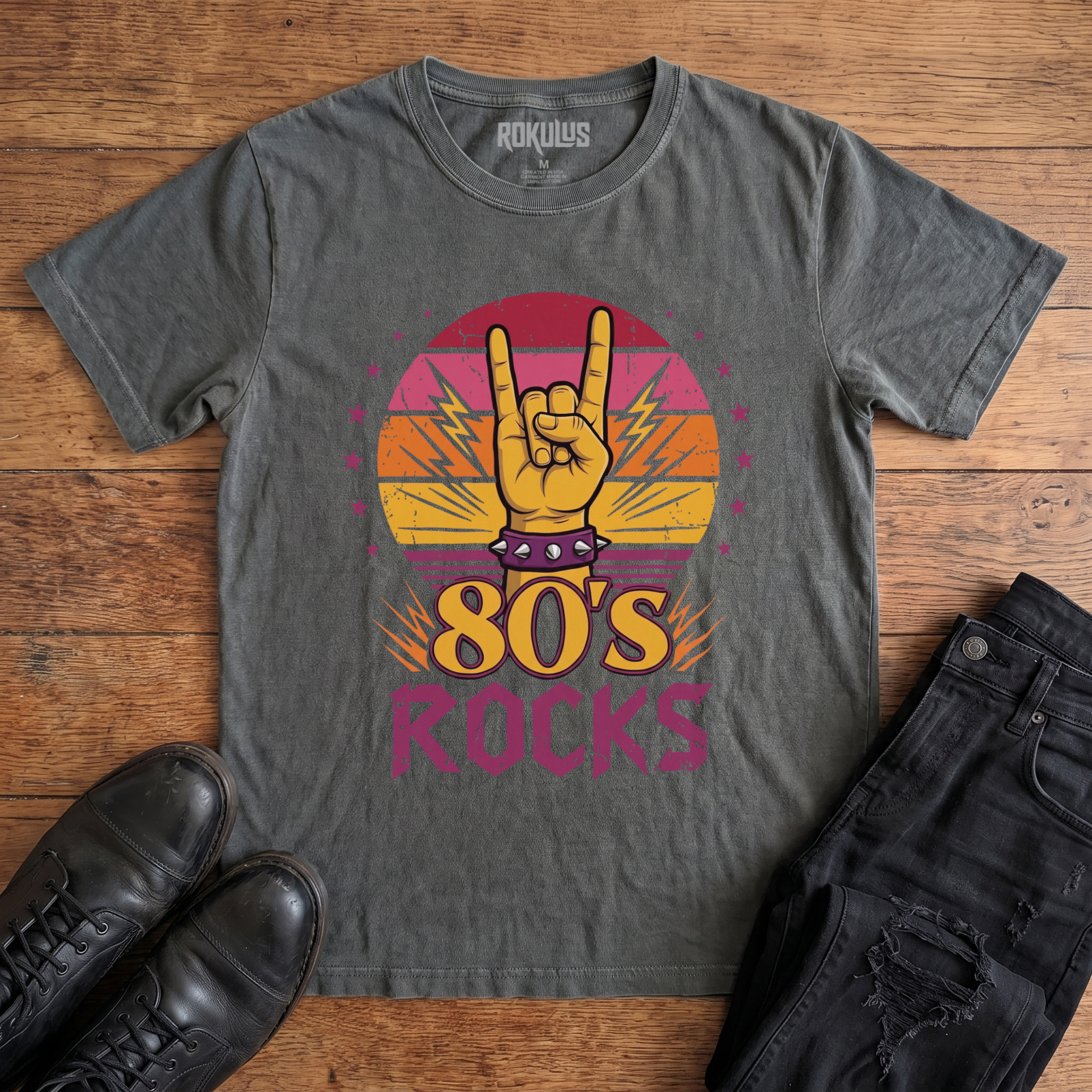 Washed charcoal Rokulus 80's Rocks vintage graphic tee laid flat on wood with black boots and jeans, front print showing a spiked-wristband horns hand against a sunset gradient circle with lightning bolts and pink stars above "80's Rocks" lettering