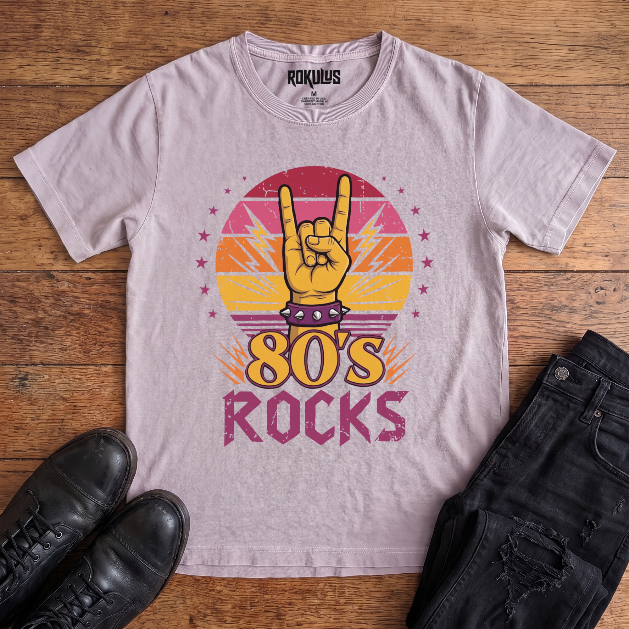 Washed lavender Rokulus 80's Rocks vintage graphic tee laid flat on wood with black boots and jeans, front print showing a spiked-wristband horns hand against a sunset gradient circle with lightning bolts and pink stars above "80's Rocks" lettering