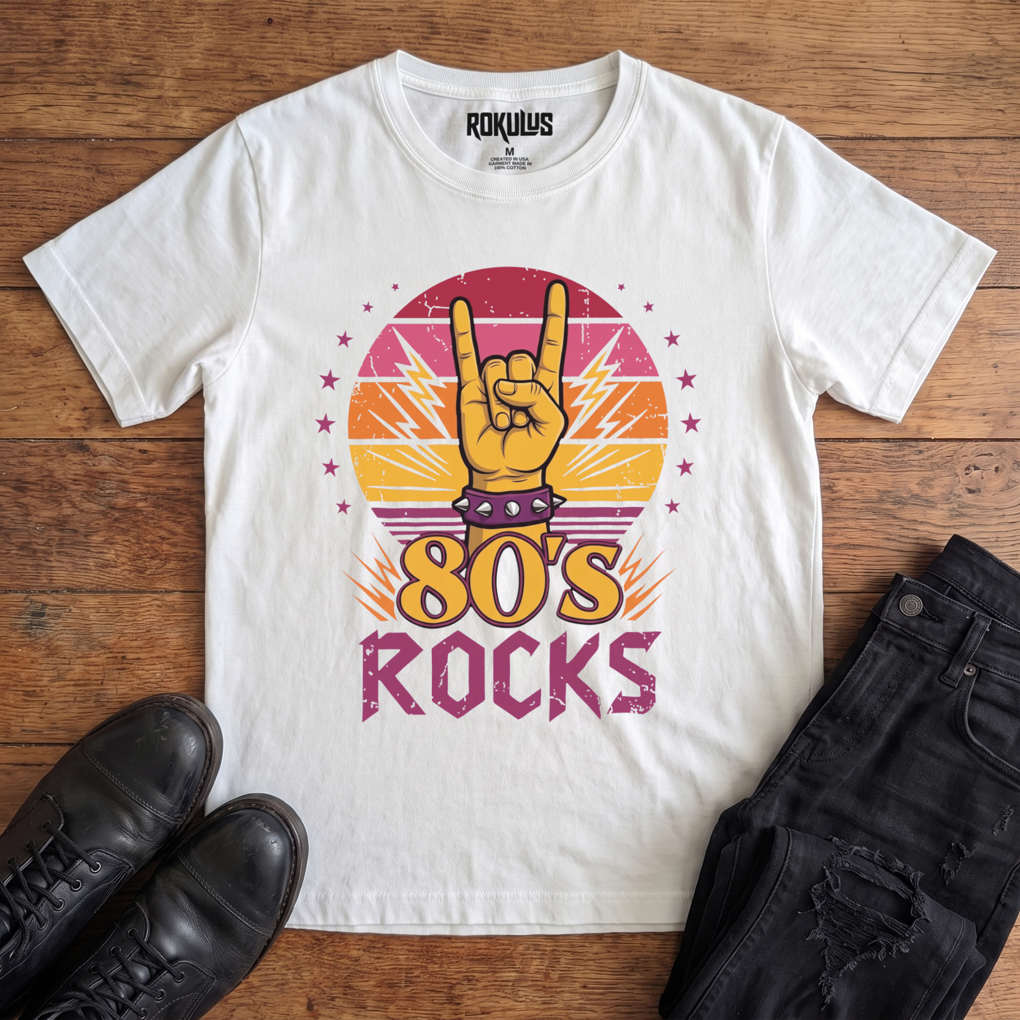 White Rokulus 80's Rocks vintage graphic tee laid flat on wood with black boots and jeans, front print showing a spiked-wristband horns hand against a sunset gradient circle with lightning bolts and pink stars above "80's Rocks" lettering