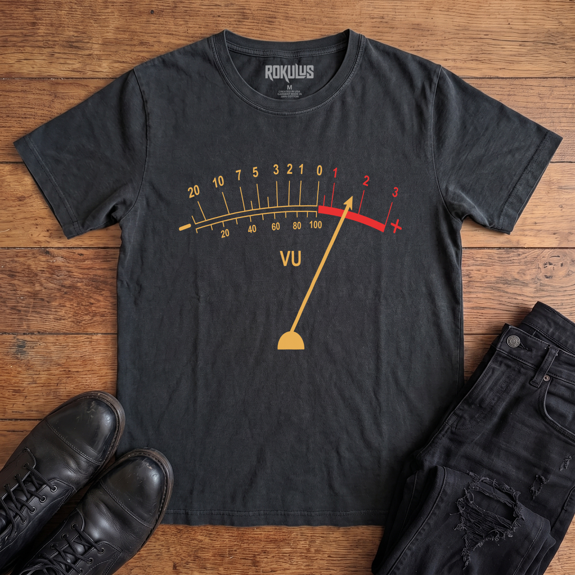Washed black Rokulus VU Meter vintage graphic tee laid flat on wood with black boots and jeans, front print showing a classic analog VU meter in golden yellow with the needle pegged into the red overload zone