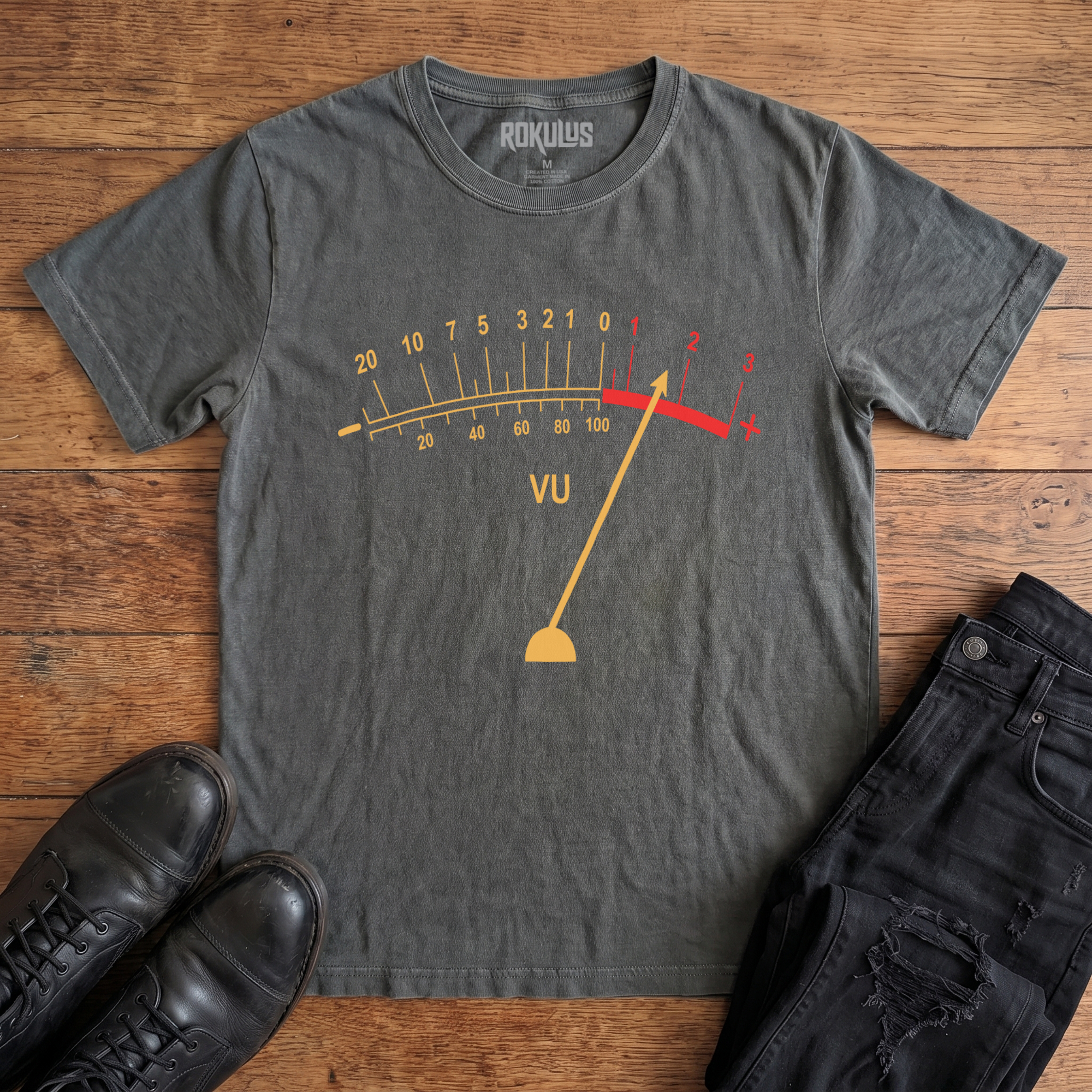 Washed charcoal Rokulus VU Meter vintage graphic tee laid flat on wood with black boots and jeans, front print showing a classic analog VU meter in golden yellow with the needle pegged into the red overload zone