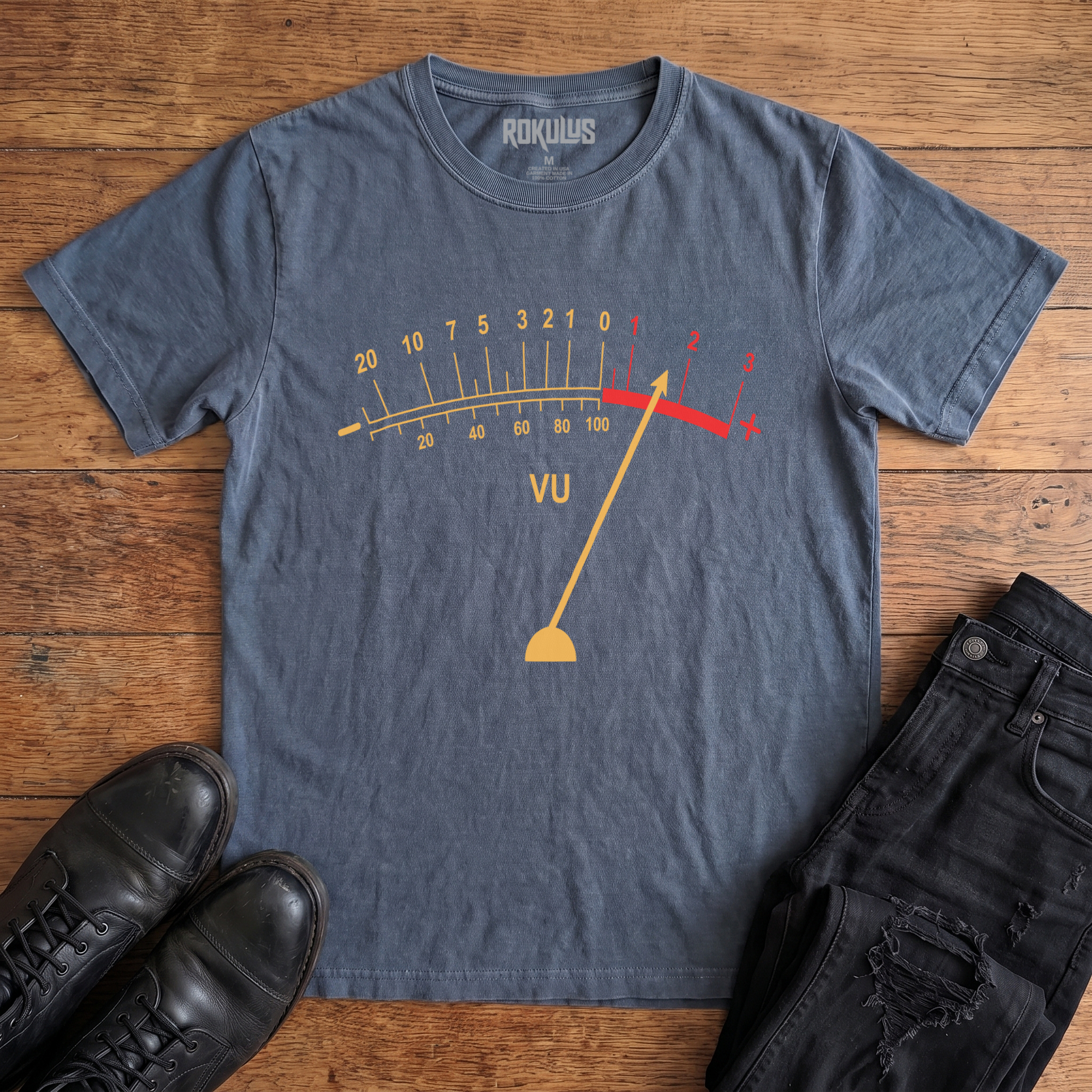 Washed navy Rokulus VU Meter vintage graphic tee laid flat on wood with black boots and jeans, front print showing a classic analog VU meter in golden yellow with the needle pegged into the red overload zone