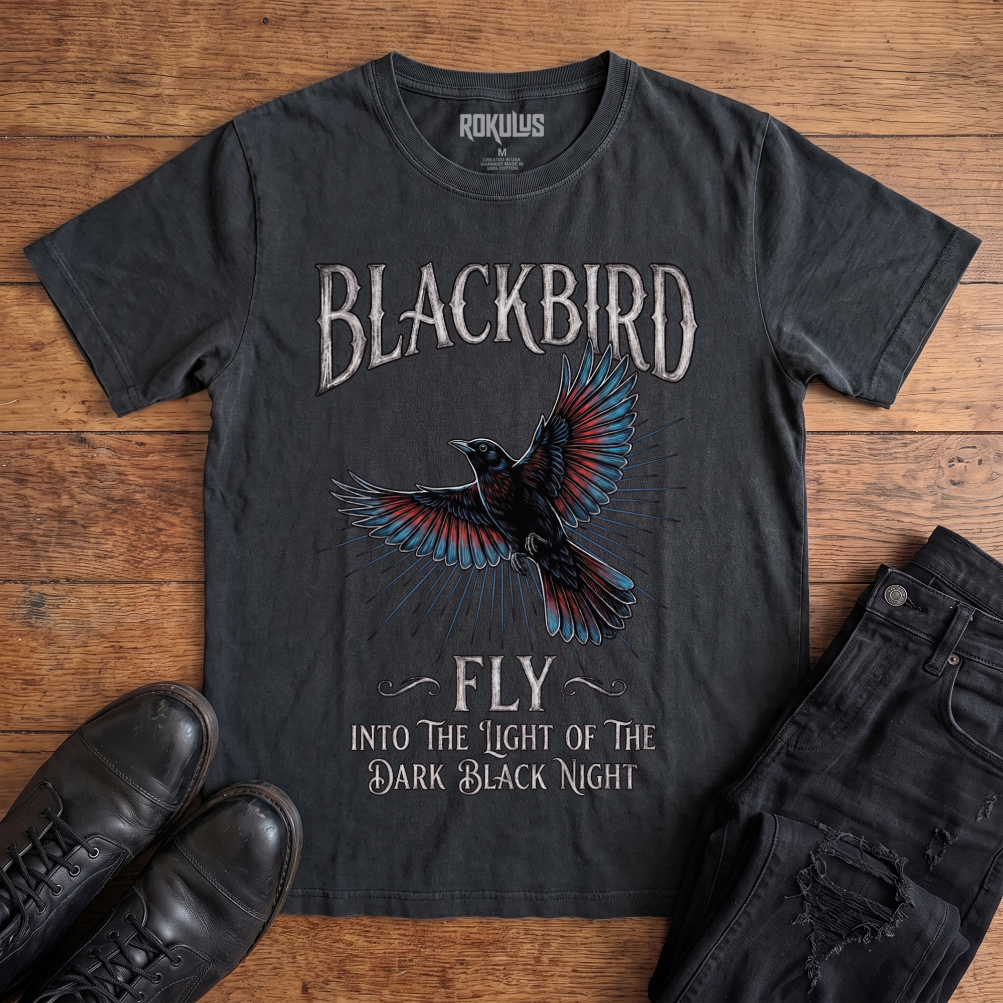 Washed black Rokulus Blackbird vintage graphic tee laid flat on wood with black boots and jeans, front print showing an inked blackbird in flight with teal and red wings and gothic "Blackbird Fly" hand-lettering