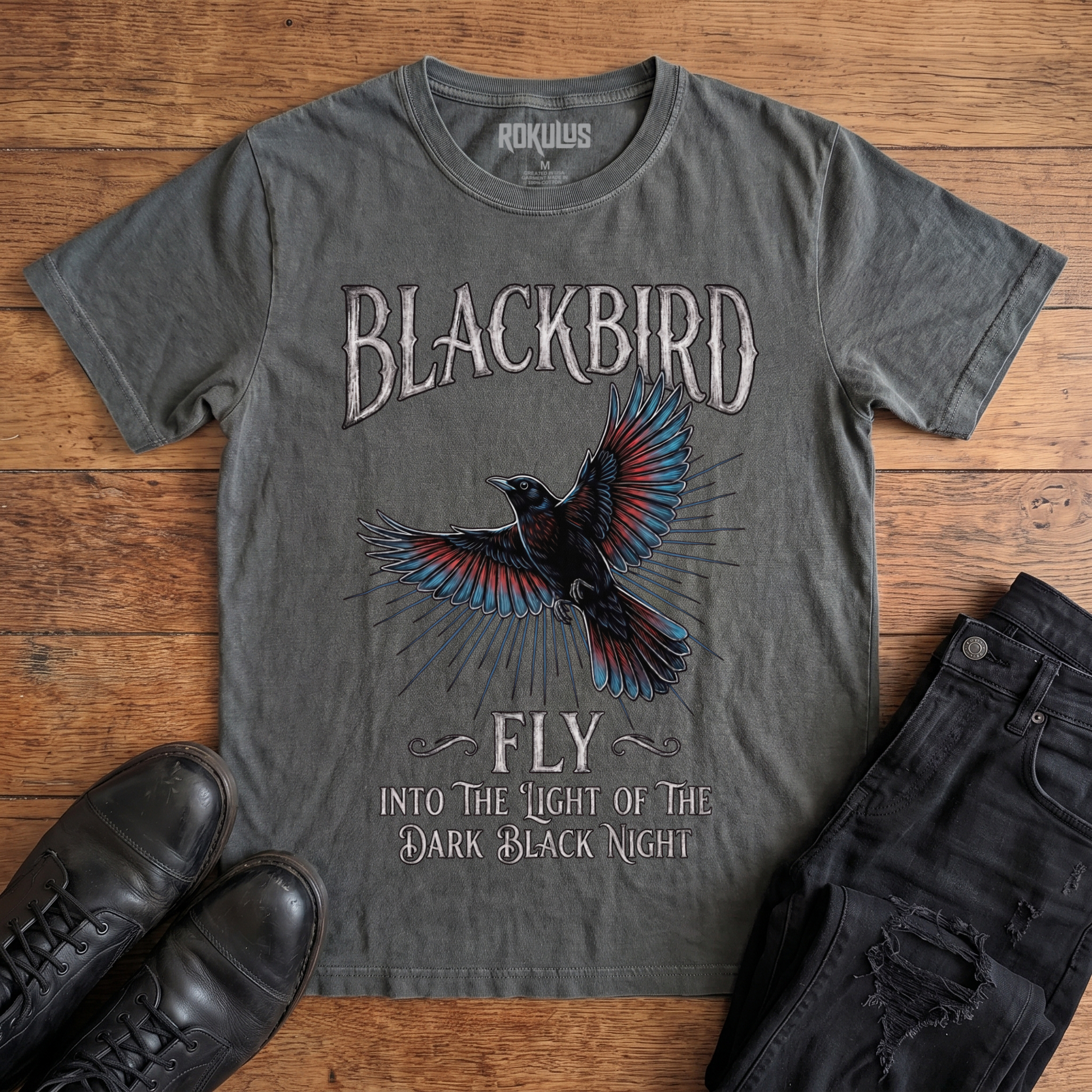 Washed charcoal Rokulus Blackbird vintage graphic tee laid flat on wood with black boots and jeans, front print showing an inked blackbird in flight with teal and red wings and gothic "Blackbird Fly" hand-lettering