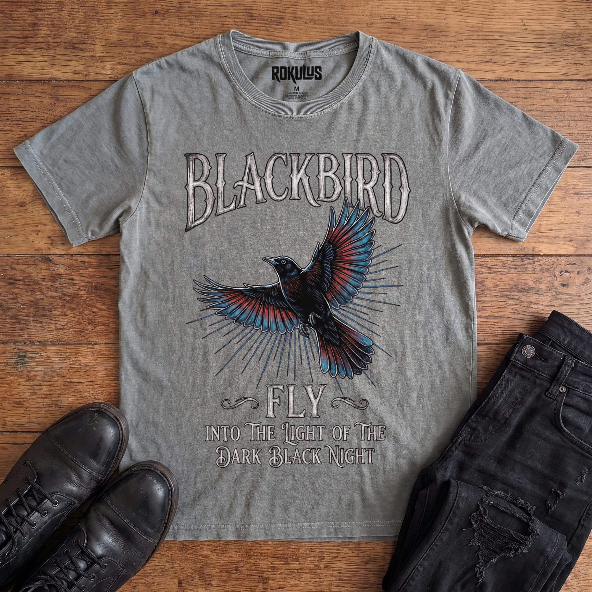 Washed grey Rokulus Blackbird vintage graphic tee laid flat on wood with black boots and jeans, front print showing an inked blackbird in flight with teal and red wings and gothic "Blackbird Fly" hand-lettering