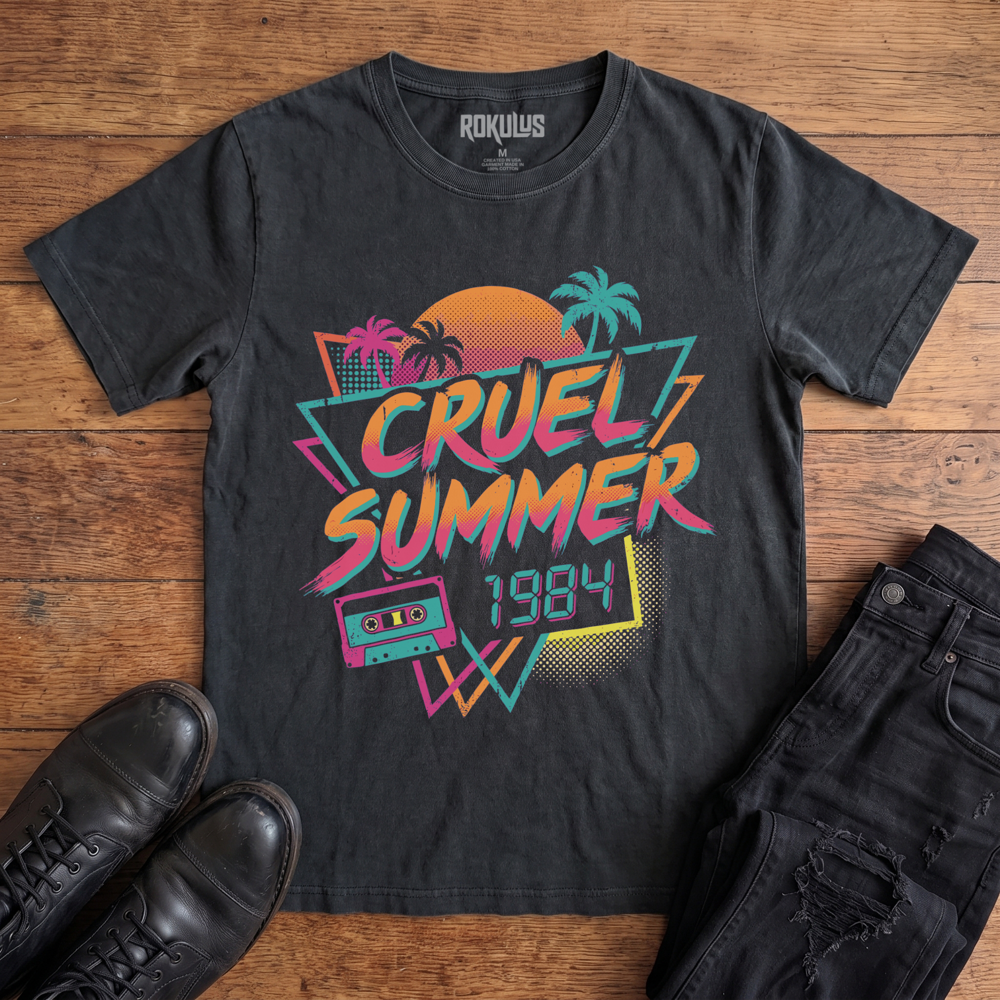 Washed black Rokulus Cruel Summer 1984 vintage graphic tee laid flat on wood with black boots and jeans, front print showing an 80s synthwave design with palm trees, a halftone sunset, and a cassette tape in neon coral, pink, teal, and yellow
