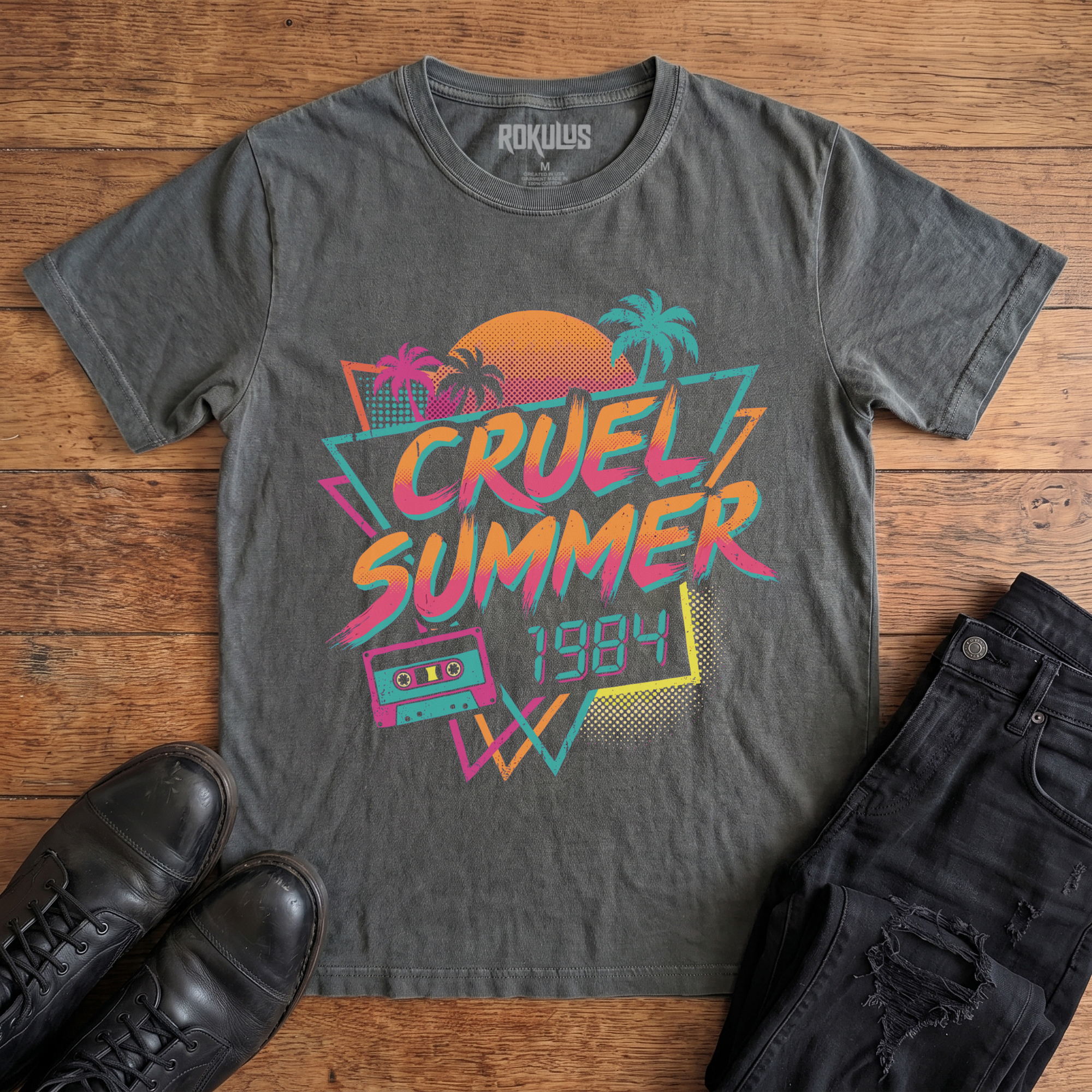 Washed charcoal Rokulus Cruel Summer 1984 vintage graphic tee laid flat on wood with black boots and jeans, front print showing an 80s synthwave design with palm trees, a halftone sunset, and a cassette tape in neon coral, pink, teal, and yellow