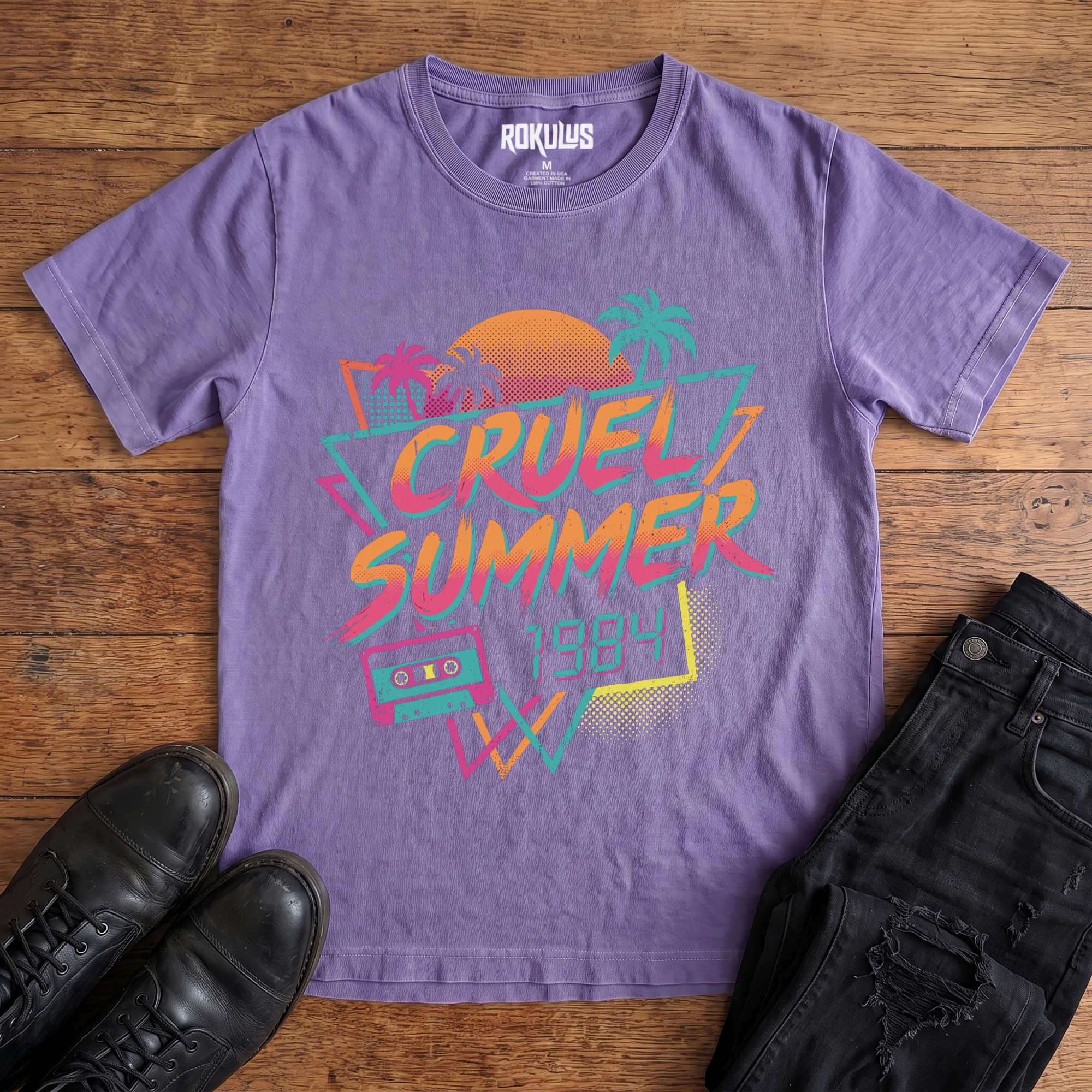 Washed purple Rokulus Cruel Summer 1984 vintage graphic tee laid flat on wood with black boots and jeans, front print showing an 80s synthwave design with palm trees, a halftone sunset, and a cassette tape in neon coral, pink, teal, and yellow
