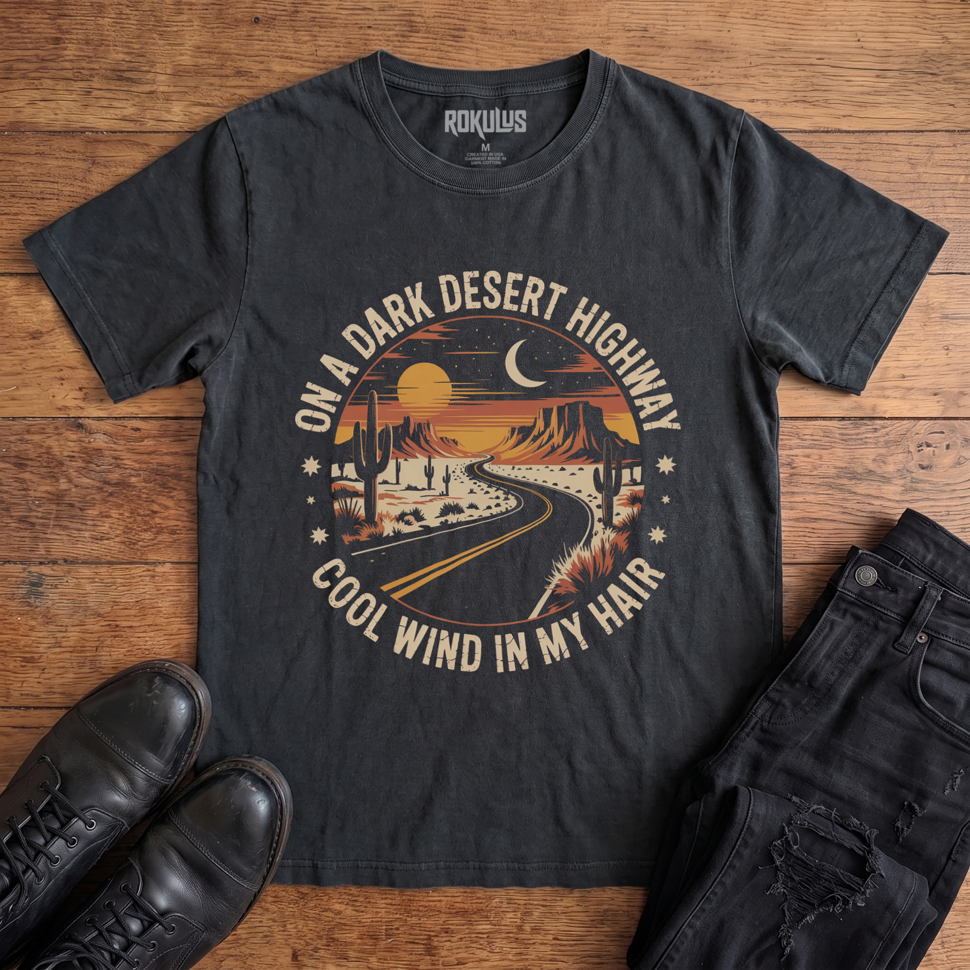 Washed black Rokulus Dark Desert Highway vintage graphic tee laid flat on wood with black boots and jeans, front print showing a circular retro desert scene with a winding highway, cacti, mesas, and a crescent moon in warm sunset tones with arched lettering