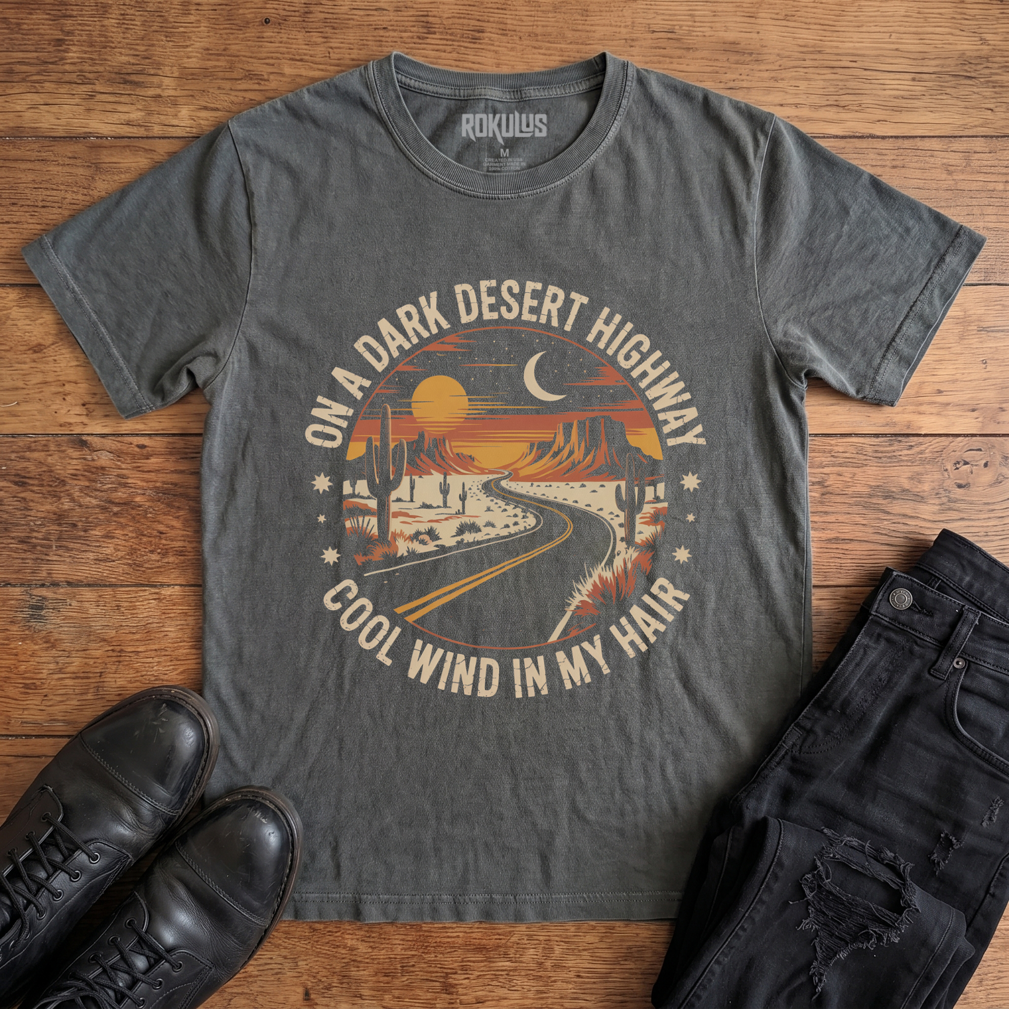 Washed charcoal Rokulus Dark Desert Highway vintage graphic tee laid flat on wood with black boots and jeans, front print showing a circular retro desert scene with a winding highway, cacti, mesas, and a crescent moon in warm sunset tones with arched letteringark Desert Highway Black Vintage Wash T-Shirt