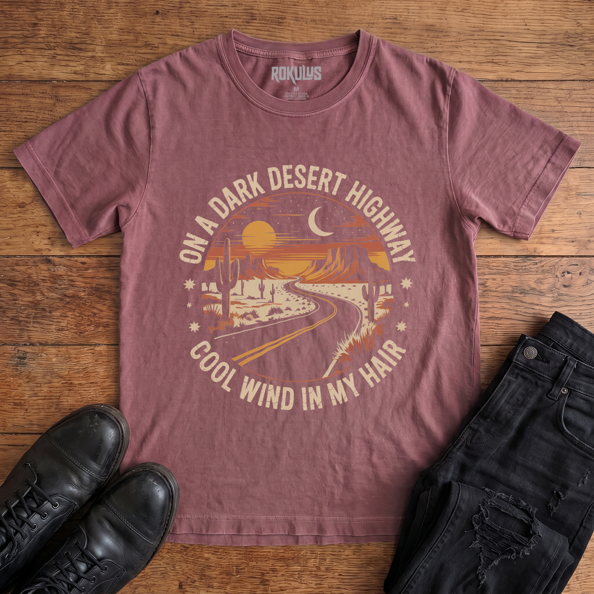 Washed maroon Rokulus Dark Desert Highway vintage graphic tee laid flat on wood with black boots and jeans, front print showing a circular retro desert scene with a winding highway, cacti, mesas, and a crescent moon in warm sunset tones with arched lettering
