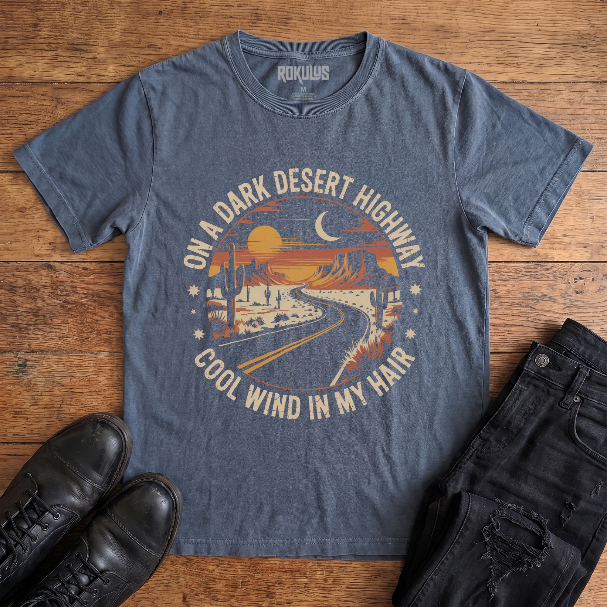 Washed navy Rokulus Dark Desert Highway vintage graphic tee laid flat on wood with black boots and jeans, front print showing a circular retro desert scene with a winding highway, cacti, mesas, and a crescent moon in warm sunset tones with arched lettering