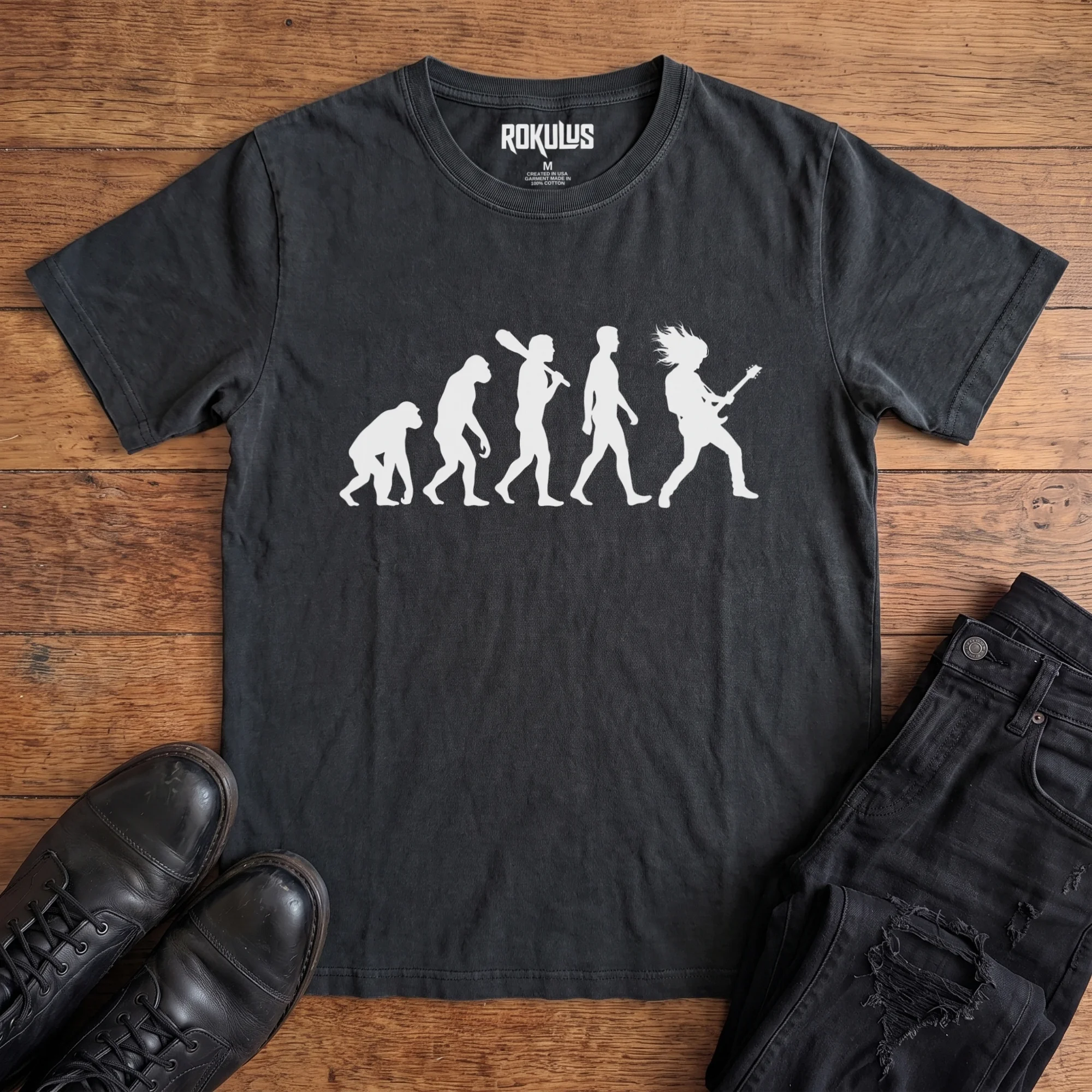 Washed black Rokulus Rock Evolution vintage graphic tee laid flat on wood with black boots and jeans, front print showing the evolution chart silhouette ending in a long-haired rocker shredding an electric guitar