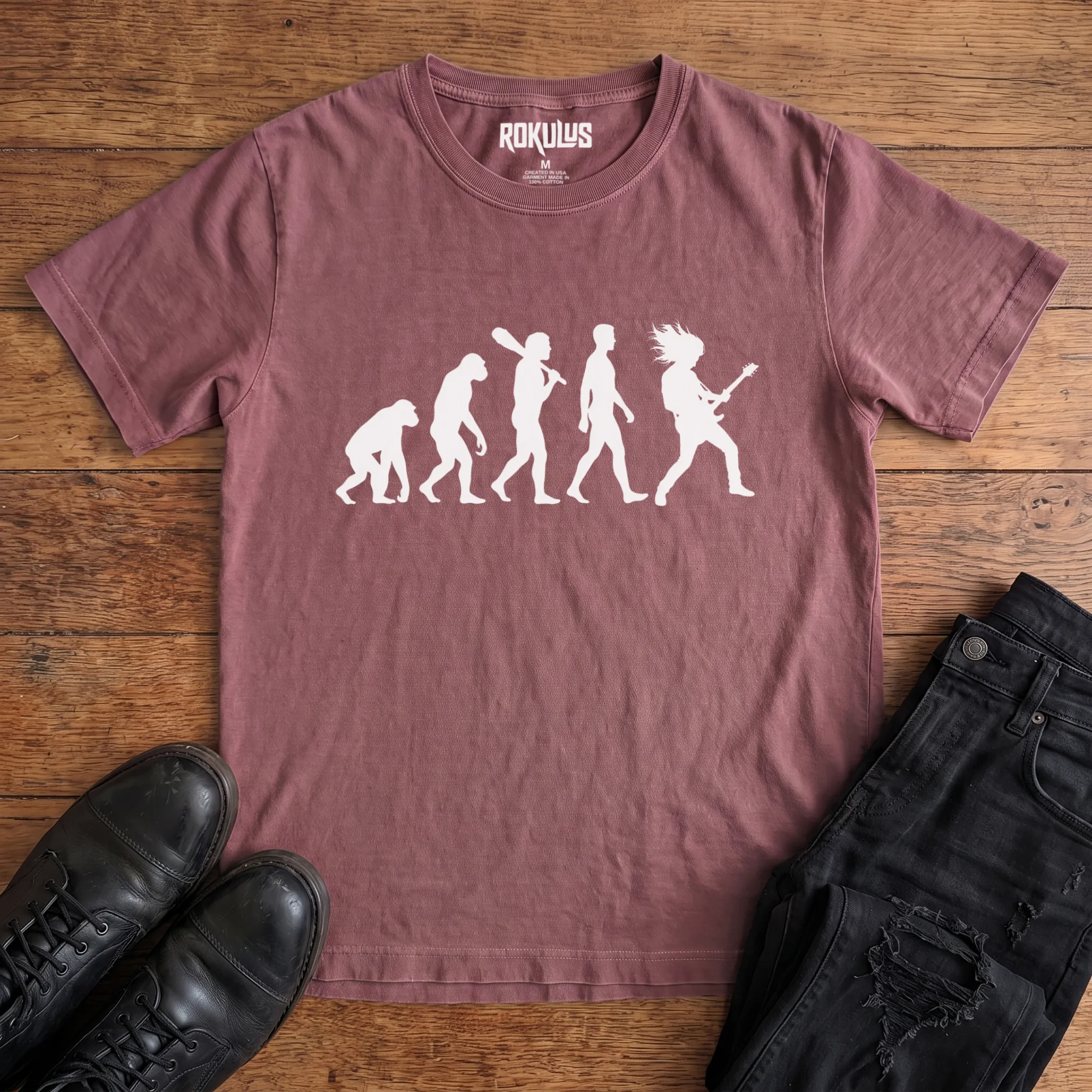 Washed maroon Rokulus Rock Evolution vintage graphic tee laid flat on wood with black boots and jeans, front print showing the evolution chart silhouette ending in a long-haired rocker shredding an electric guitar