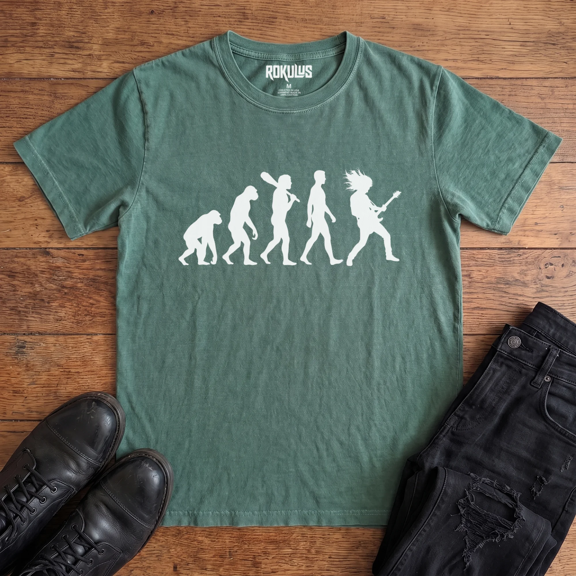Washed pine Rokulus Rock Evolution vintage graphic tee laid flat on wood with black boots and jeans, front print showing the evolution chart silhouette ending in a long-haired rocker shredding an electric guitar