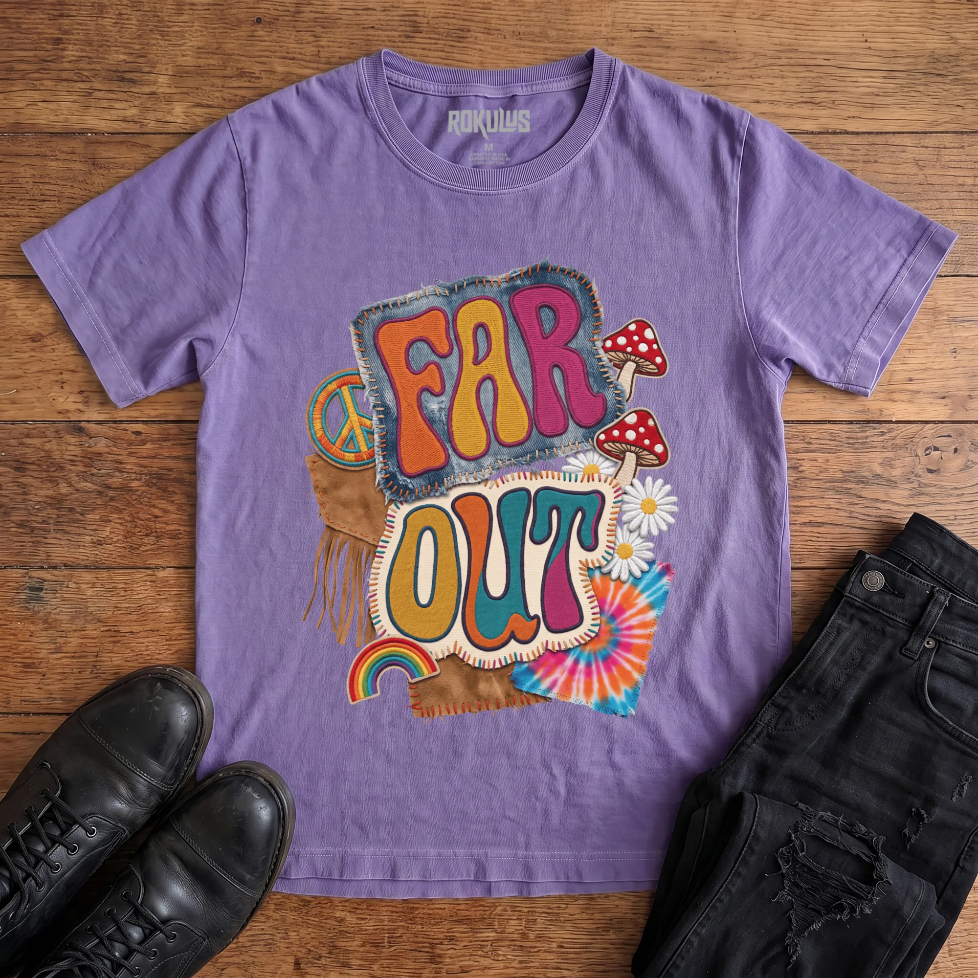 Washed purple Rokulus Far Out vintage graphic tee laid flat on wood with black boots and jeans, front print showing a groovy 70s patch collage with mushrooms, daisies, a peace sign, rainbow, and tie-dye around puffy “Far Out” lettering
