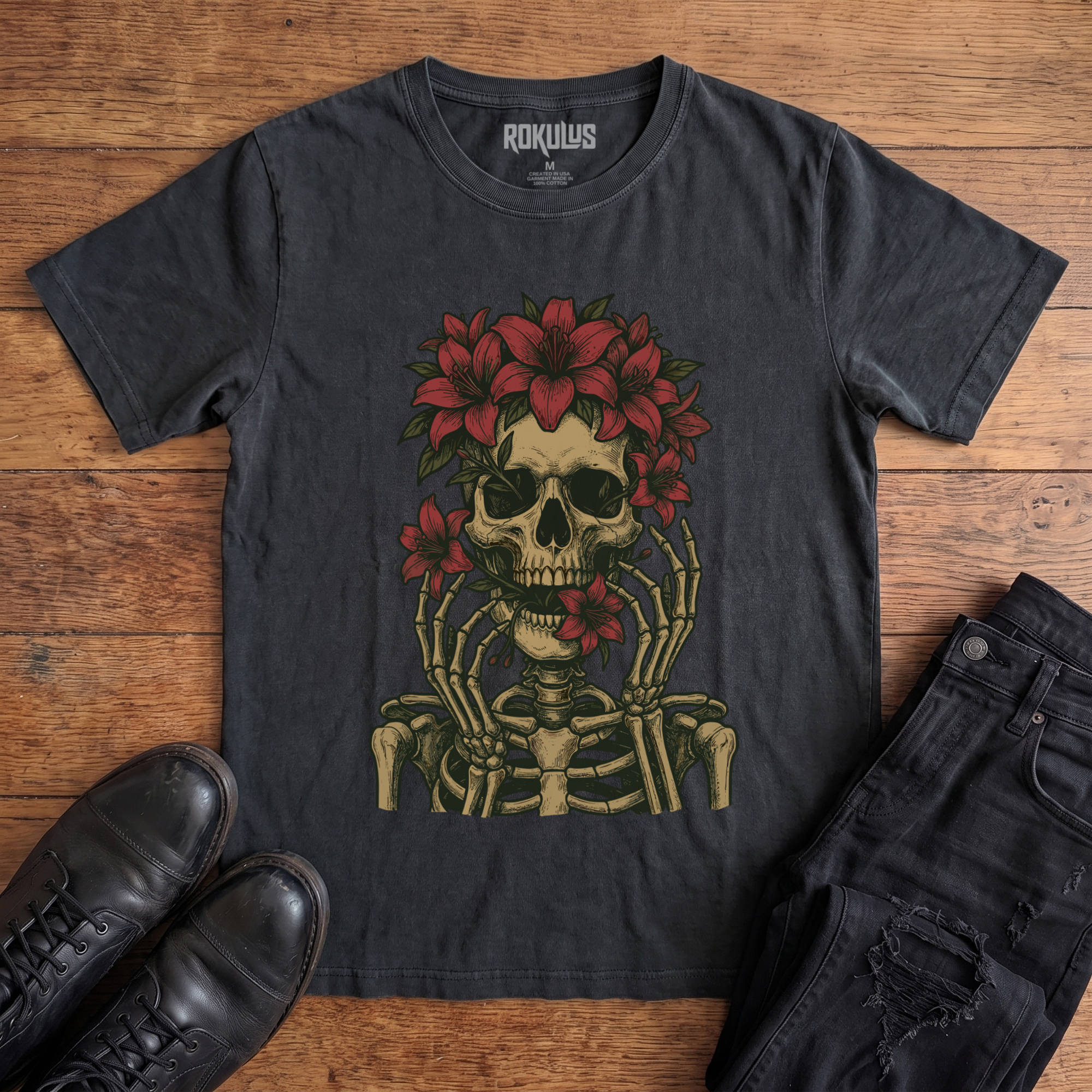 Washed black Rokulus Floral Skull vintage graphic tee laid flat on wood with black boots and jeans, front print showing a skeleton crowned with red lilies and skeletal hands framing the face in fine tattoo-flash line work