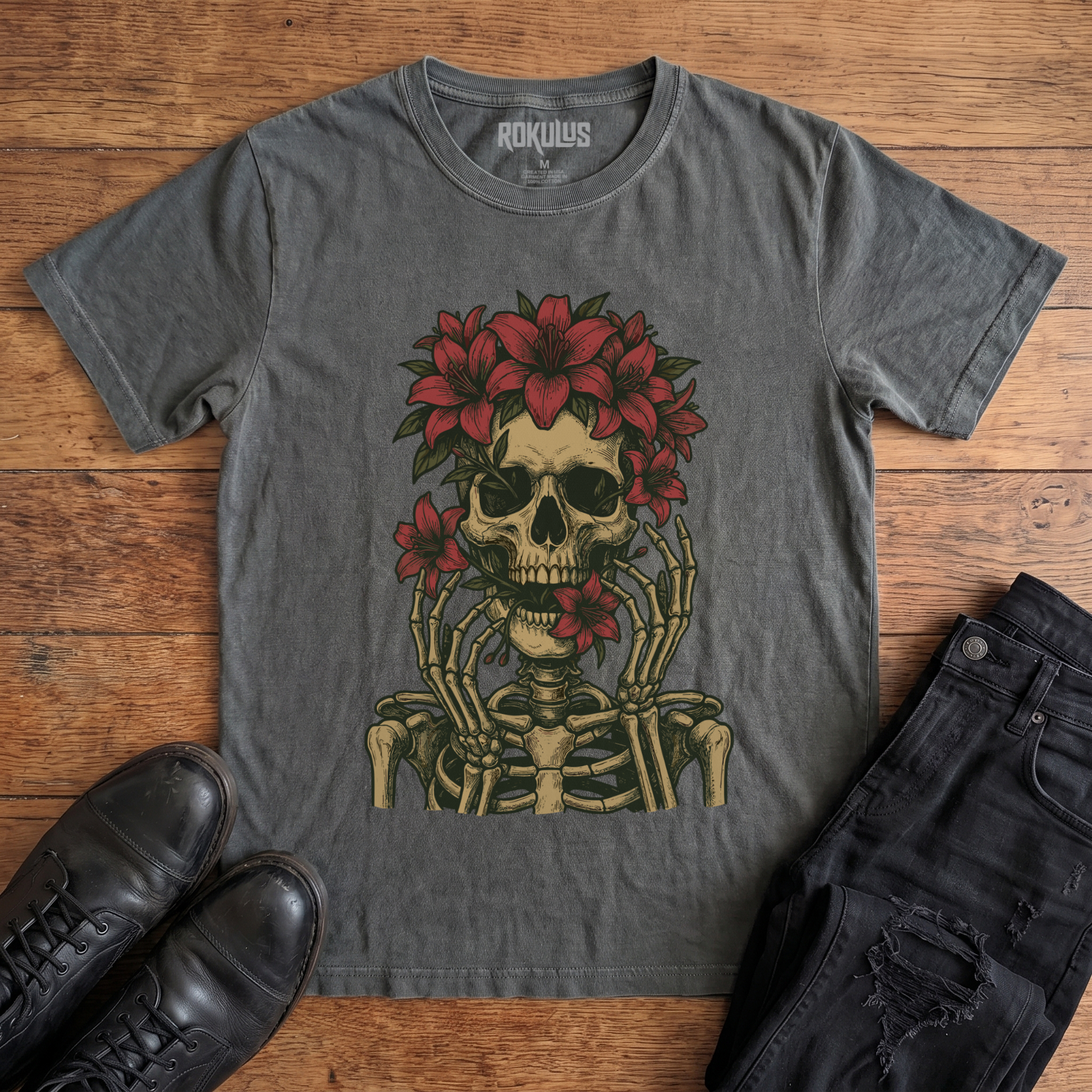 Washed charcoal Rokulus Floral Skull vintage graphic tee laid flat on wood with black boots and jeans, front print showing a skeleton crowned with red lilies and skeletal hands framing the face in fine tattoo-flash line work