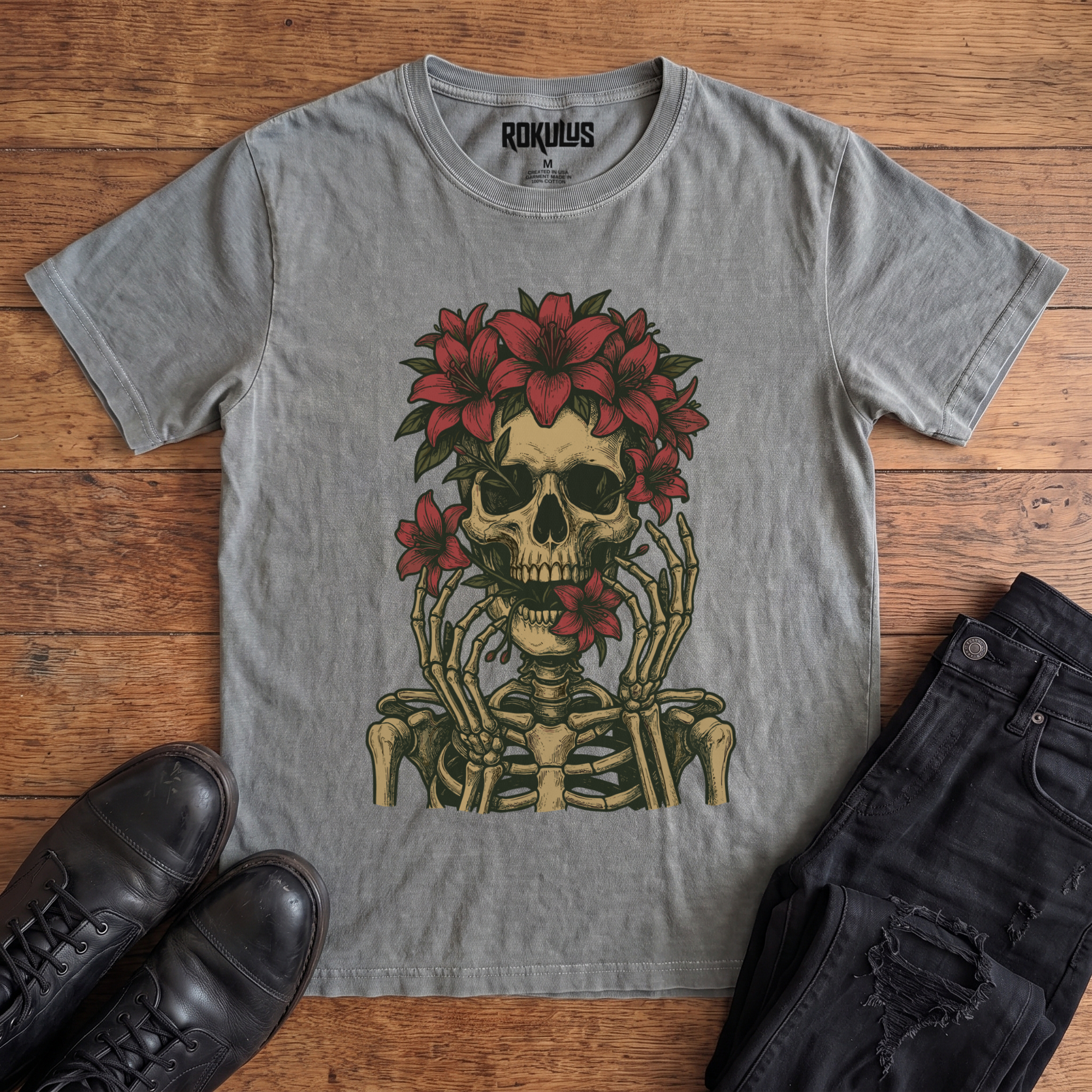 Washed grey Rokulus Floral Skull vintage graphic tee laid flat on wood with black boots and jeans, front print showing a skeleton crowned with red lilies and skeletal hands framing the face in fine tattoo-flash line work
