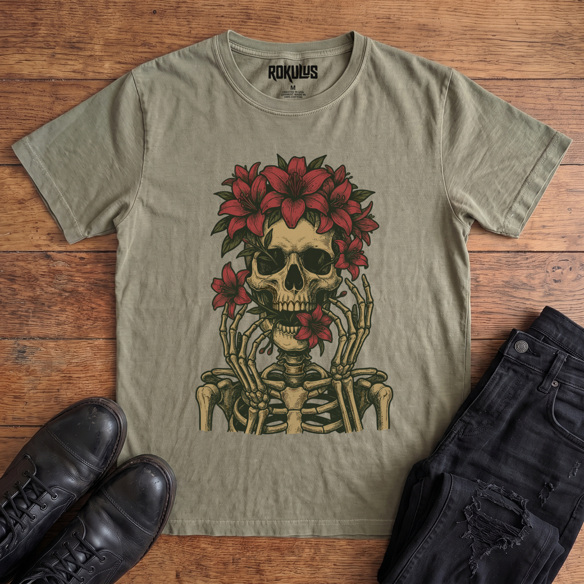 Washed olive green Rokulus Floral Skull vintage graphic tee laid flat on wood with black boots and jeans, front print showing a skeleton crowned with red lilies and skeletal hands framing the face in fine tattoo-flash line work