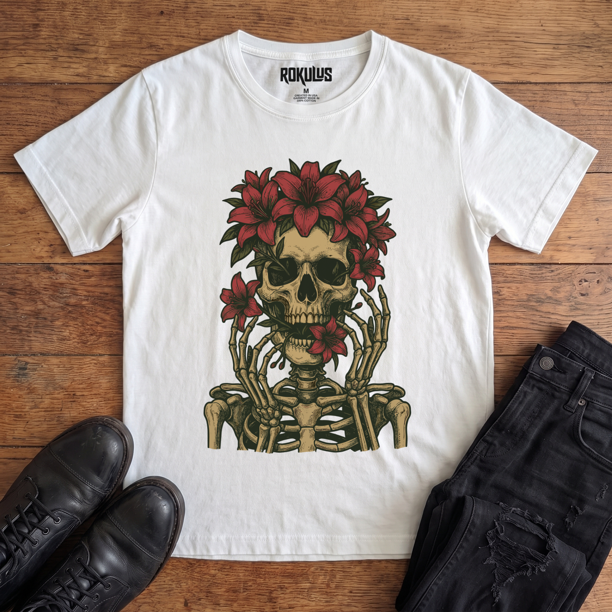 White Rokulus Floral Skull vintage graphic tee laid flat on wood with black boots and jeans, front print showing a skeleton crowned with red lilies and skeletal hands framing the face in fine tattoo-flash line work