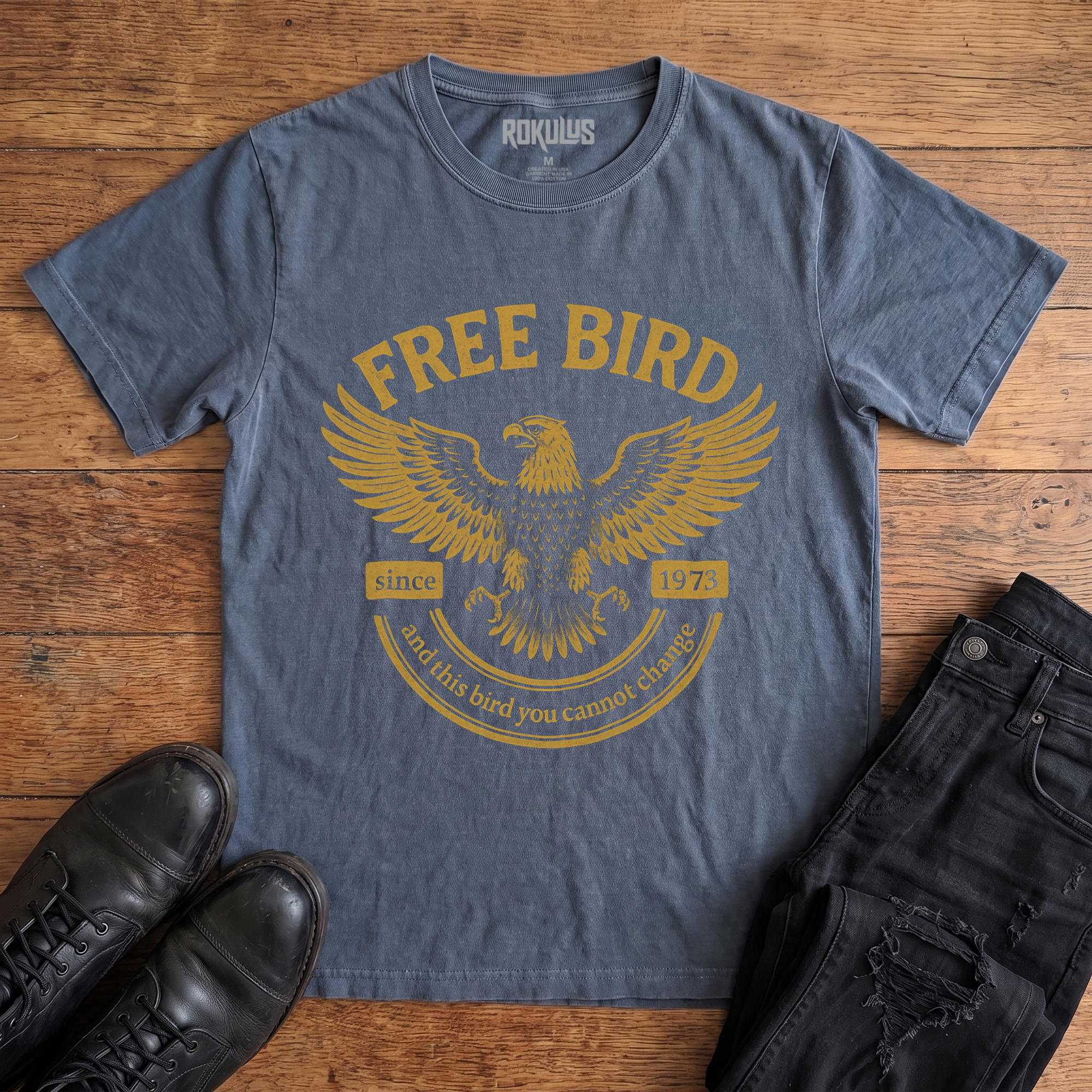Washed navy blue Rokulus Free Bird vintage graphic tee laid flat on wood with black boots and jeans, front print showing a spread-winged eagle in distressed gold with arched "Free Bird" lettering and vintage banner type