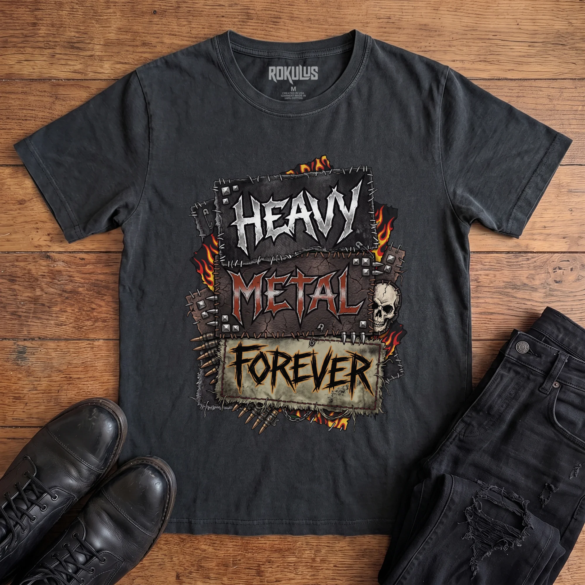 Washed black Rokulus Heavy Metal Forever vintage graphic tee laid flat on wood with black boots and jeans, front print showing a battle-vest design with studs, flames, bullets, a skull, and chrome "Heavy Metal Forever" lettering across stitched patches
