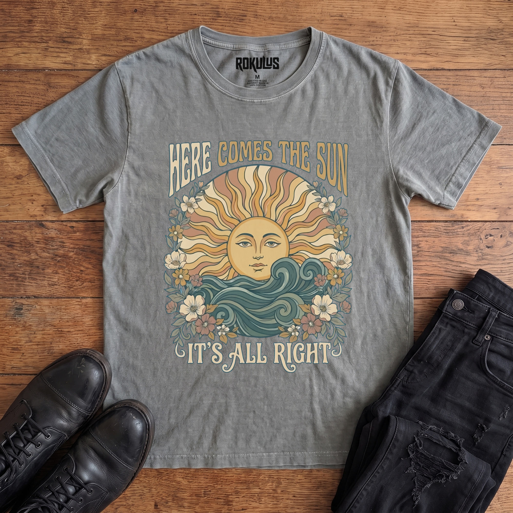 Washed grey Rokulus Here Comes The Sun vintage graphic tee laid flat on wood with black boots and jeans, front print showing a boho celestial sun face over ocean waves framed by wildflowers with groovy "Here Comes The Sun It's All Right" lettering