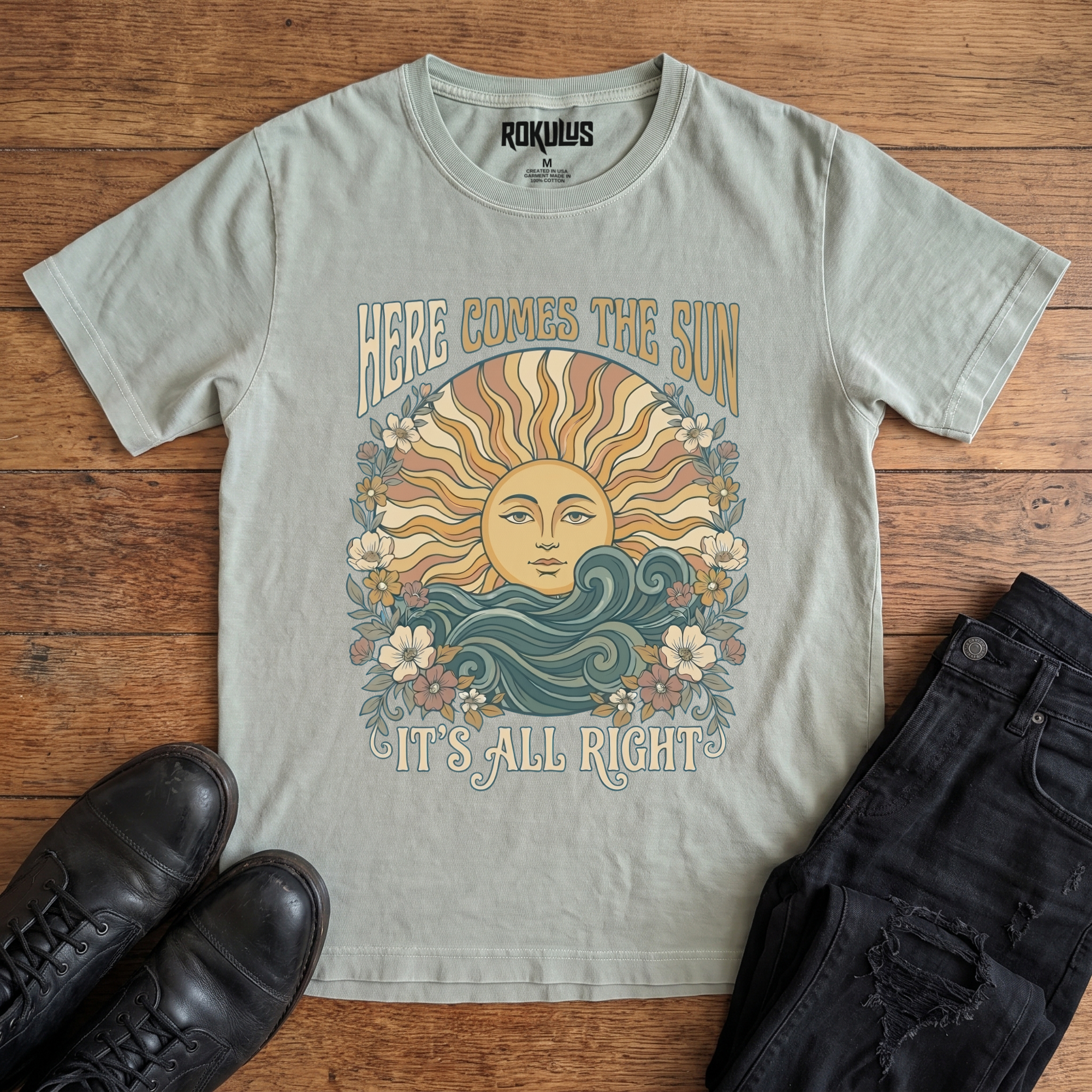 Washed thyme Rokulus Here Comes The Sun vintage graphic tee laid flat on wood with black boots and jeans, front print showing a boho celestial sun face over ocean waves framed by wildflowers with groovy "Here Comes The Sun It's All Right" lettering