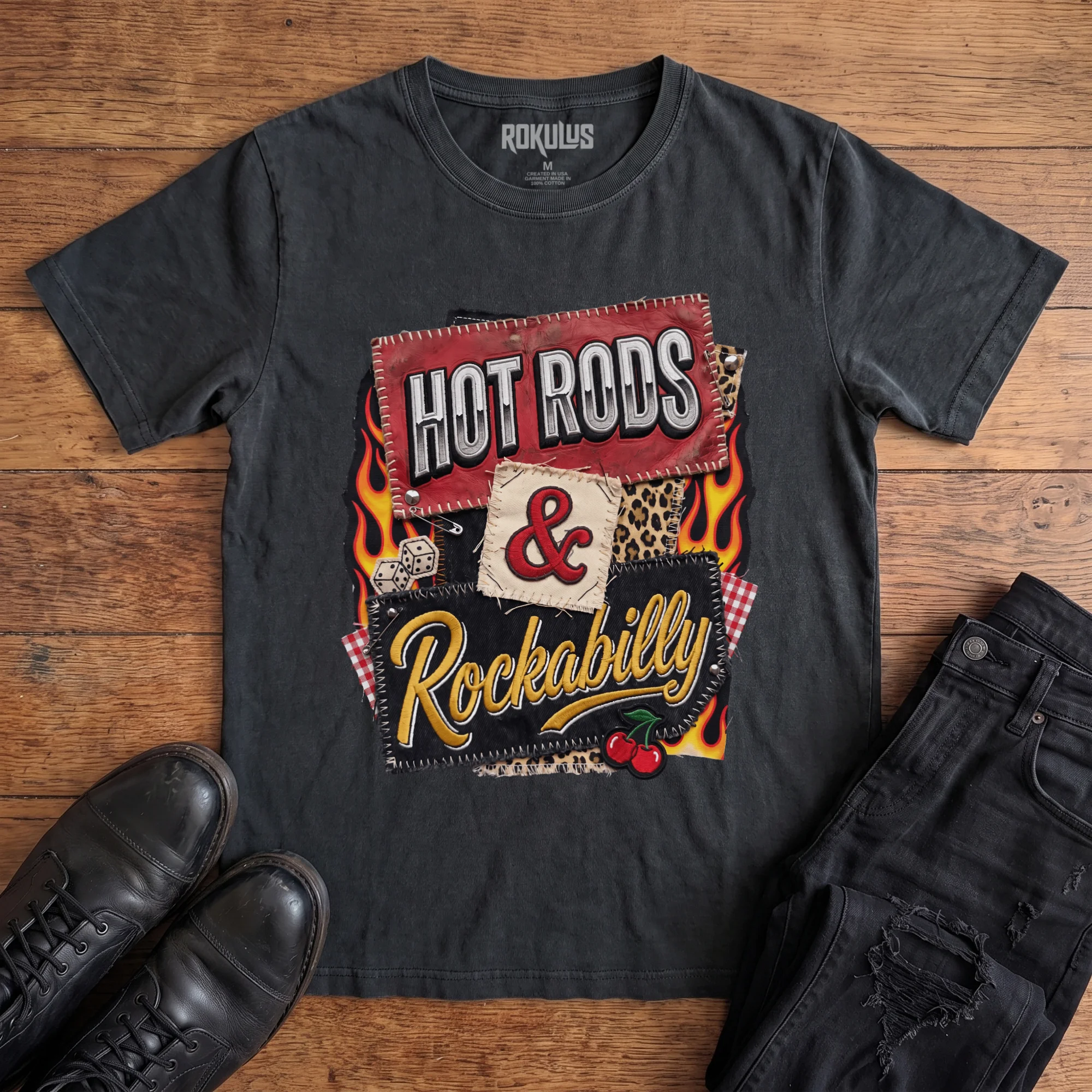 Washed black Rokulus Hot Rods & Rockabilly vintage graphic tee laid flat on wood with black boots and jeans, front print showing a patchwork kustom kulture design with flames, dice, cherries, and leopard print under stitched "Hot Rods & Rockabilly" lettering