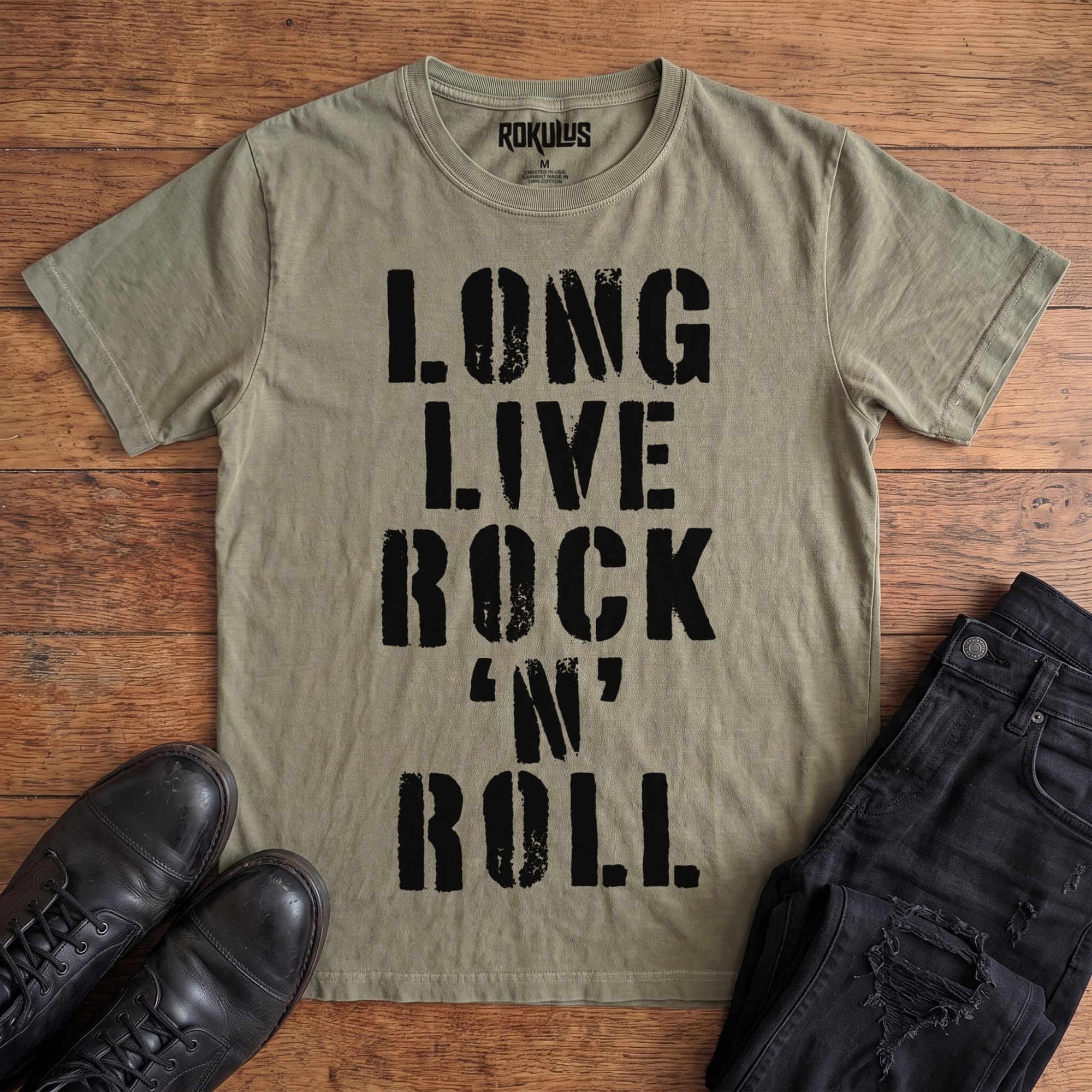 Washed olive Rokulus Long Live Rock 'n' Roll vintage graphic tee laid flat on wood with black boots and jeans, front print showing "Long Live Rock 'n' Roll" stacked vertically in heavy distressed black stencil lettering