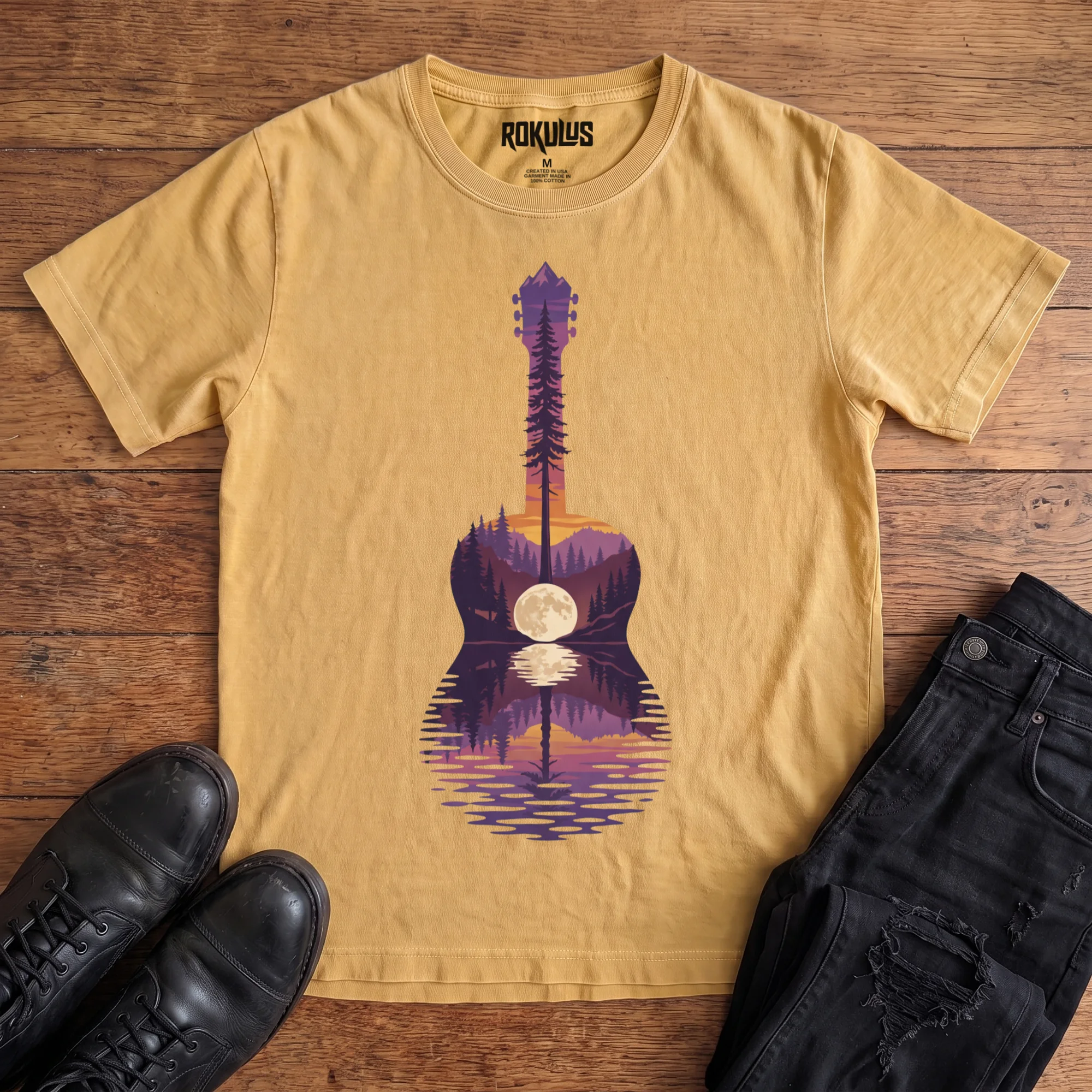 Washed Mustard Rokulus Guitar Lake vintage graphic tee laid flat on wood with black boots and jeans, front print showing an acoustic guitar formed from a pine forest, mountains, and a full moon reflecting on a lake at sunset in purple and orange