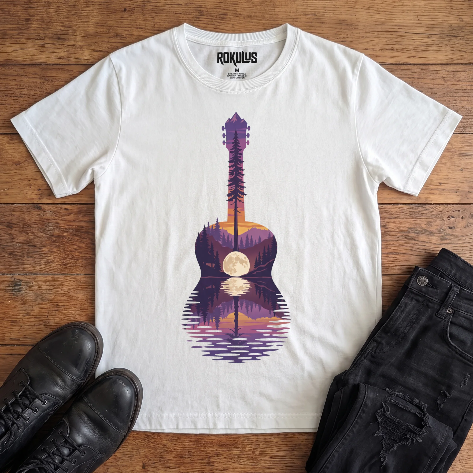 White Rokulus Guitar Lake vintage graphic tee laid flat on wood with black boots and jeans, front print showing an acoustic guitar formed from a pine forest, mountains, and a full moon reflecting on a lake at sunset in purple and orange