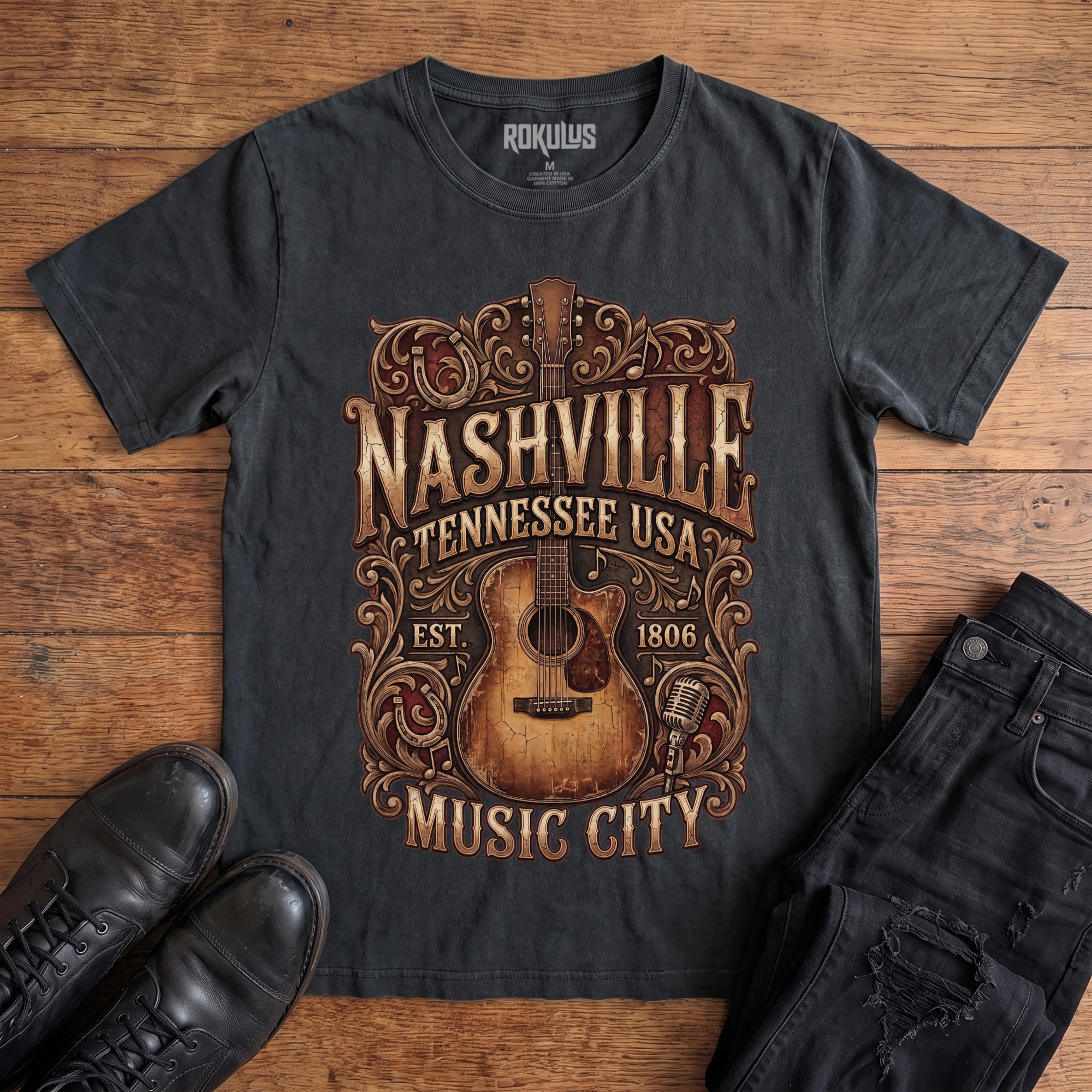 Washed black Rokulus Nashville Music City vintage graphic tee laid flat on wood with black boots and jeans, front print showing a weathered acoustic guitar with ornate scrollwork and saloon-style "Nashville Tennessee USA Music City" lettering in aged gold and brown