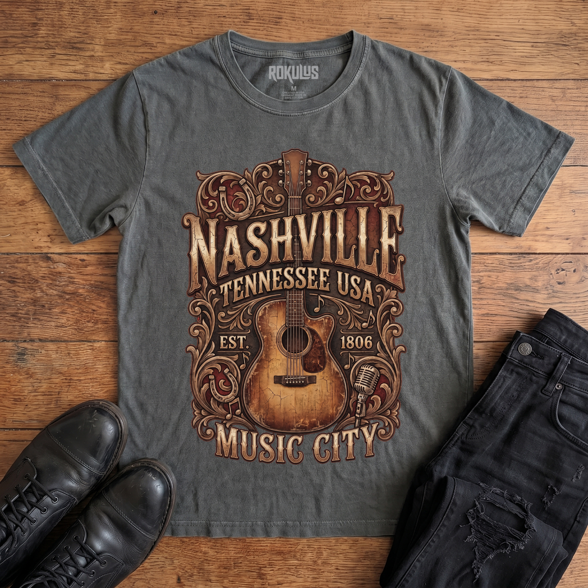 Washed charcoal Rokulus Nashville Music City vintage graphic tee laid flat on wood with black boots and jeans, front print showing a weathered acoustic guitar with ornate scrollwork and saloon-style "Nashville Tennessee USA Music City" lettering in aged gold and brown