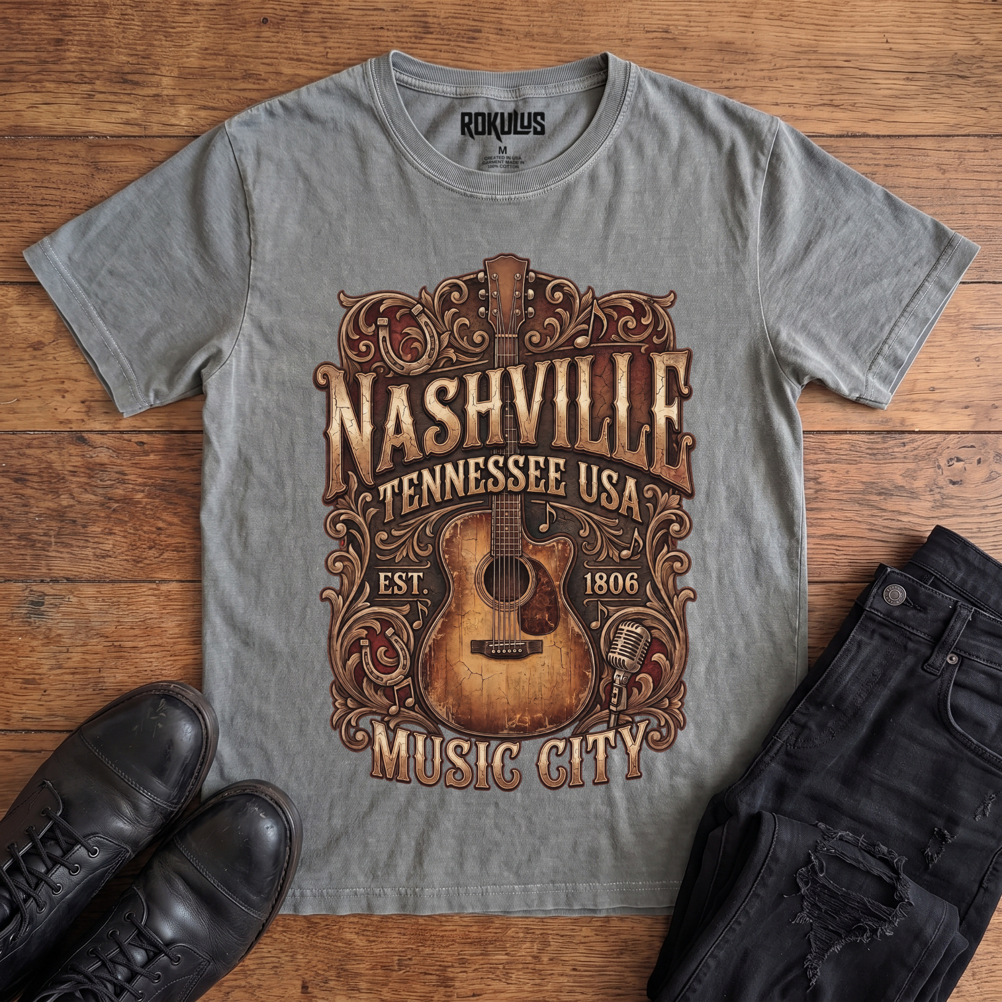 Washed grey Rokulus Nashville Music City vintage graphic tee laid flat on wood with black boots and jeans, front print showing a weathered acoustic guitar with ornate scrollwork and saloon-style "Nashville Tennessee USA Music City" lettering in aged gold and brown