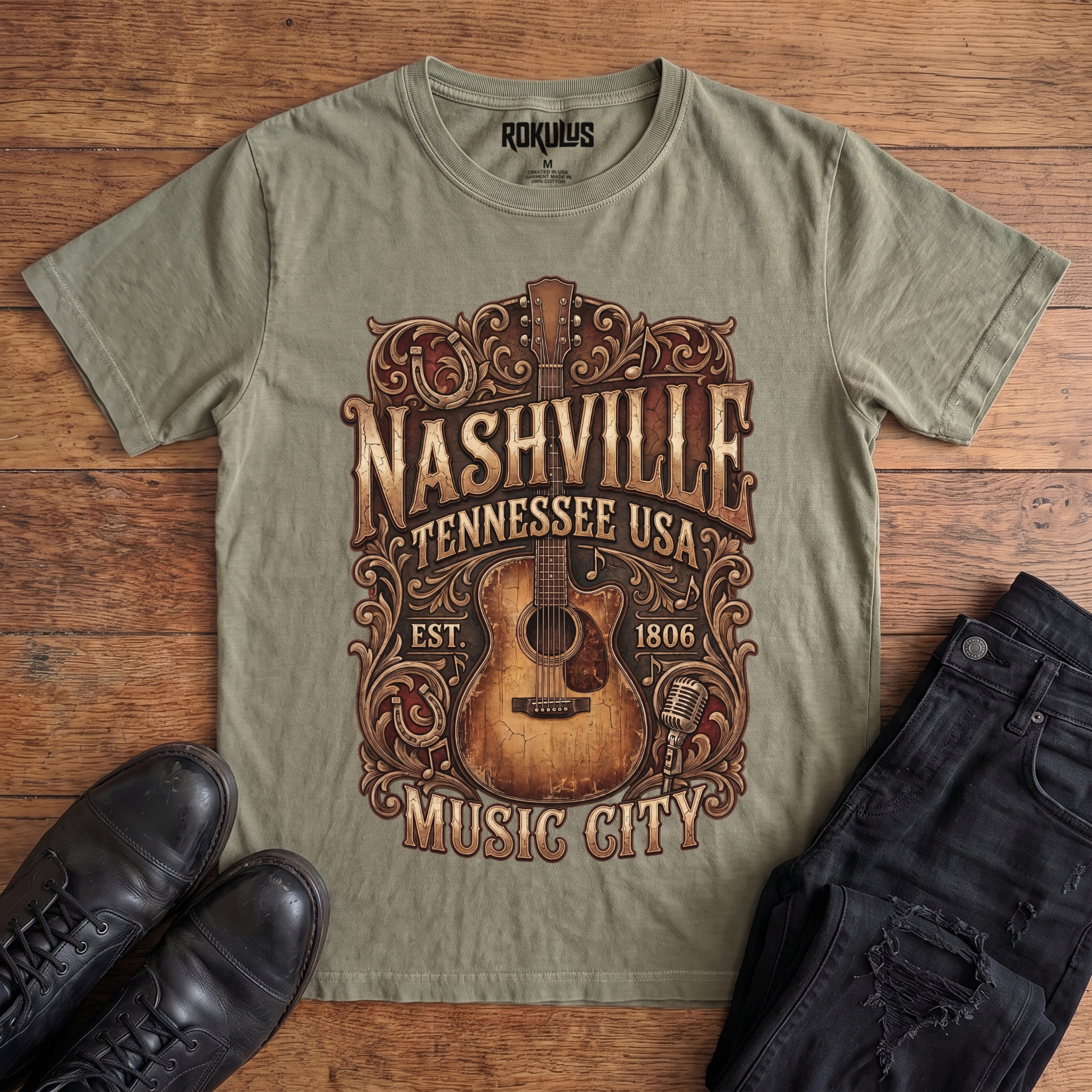 Washed olive Rokulus Nashville Music City vintage graphic tee laid flat on wood with black boots and jeans, front print showing a weathered acoustic guitar with ornate scrollwork and saloon-style "Nashville Tennessee USA Music City" lettering in aged gold and brown
