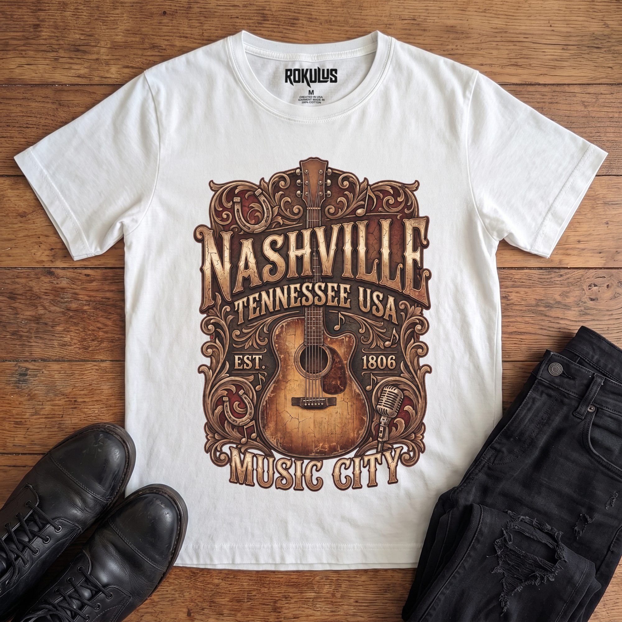 White Rokulus Nashville Music City vintage graphic tee laid flat on wood with black boots and jeans, front print showing a weathered acoustic guitar with ornate scrollwork and saloon-style "Nashville Tennessee USA Music City" lettering in aged gold and brown