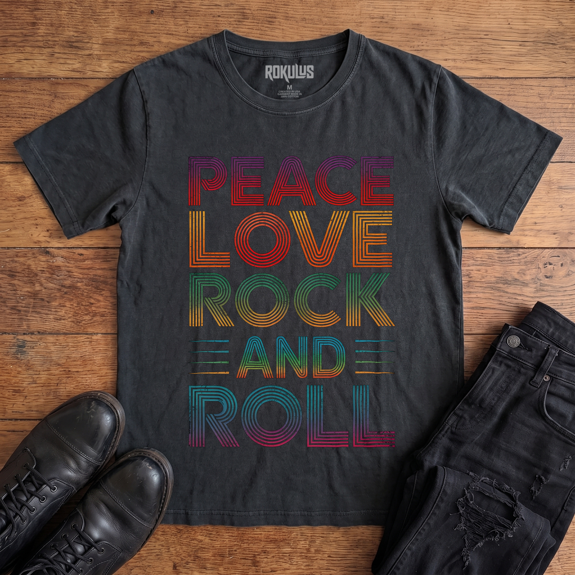 Washed black Rokulus Peace Love Rock and Roll vintage graphic tee laid flat on wood with black boots and jeans, front print showing "Peace Love Rock and Roll" stacked in five lines of layered retro type in a rainbow gradient