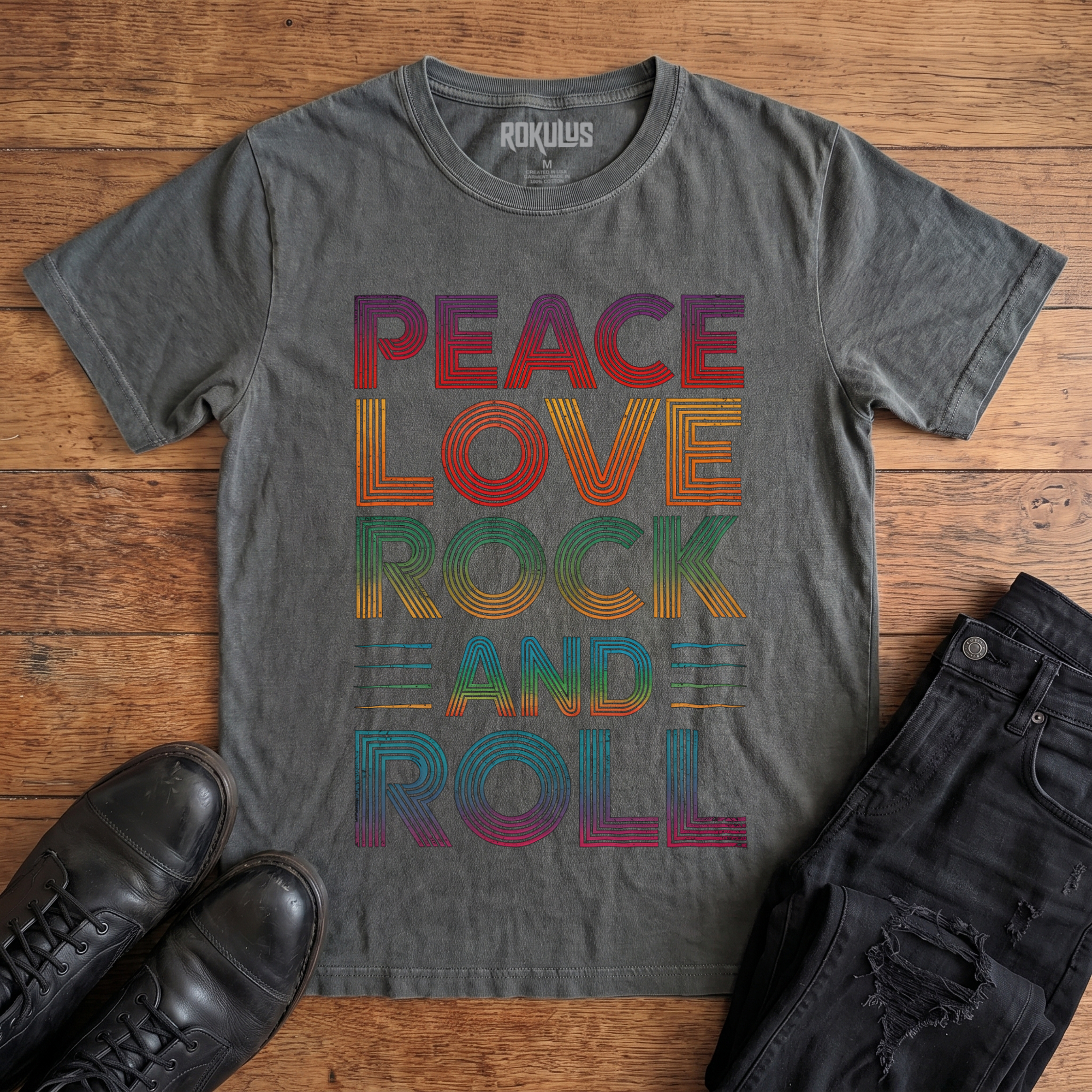 Washed charcoal Rokulus Peace Love Rock and Roll vintage graphic tee laid flat on wood with black boots and jeans, front print showing "Peace Love Rock and Roll" stacked in five lines of layered retro type in a rainbow gradient