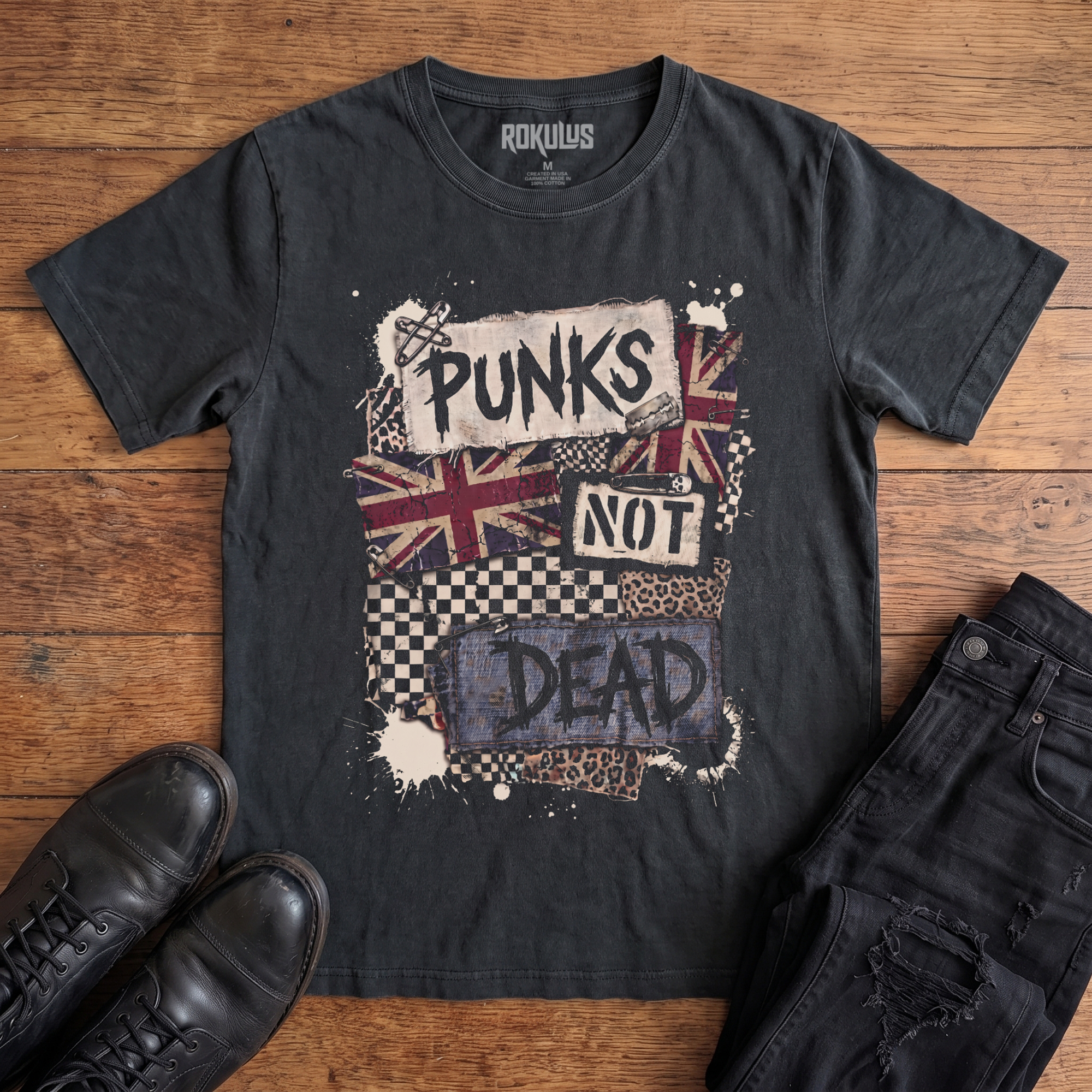 Washed black Rokulus Punks Not Dead vintage graphic tee laid flat on wood with black boots and jeans, front print showing a punk collage with ransom-style "Punks Not Dead" lettering, Union Jack, checkerboard, leopard print, and safety pins
