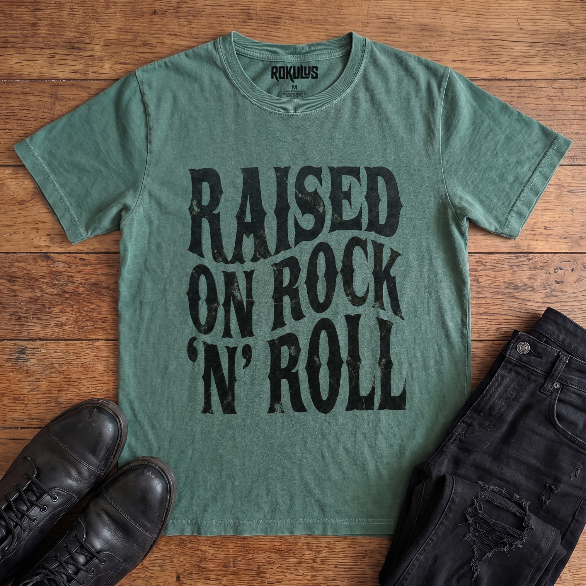 Washed pine Rokulus Raised On Rock 'N' Roll vintage graphic tee laid flat on wood with black boots and jeans, front print showing "Raised On Rock 'N' Roll" stacked in three lines of heavy wavy distressed black lettering