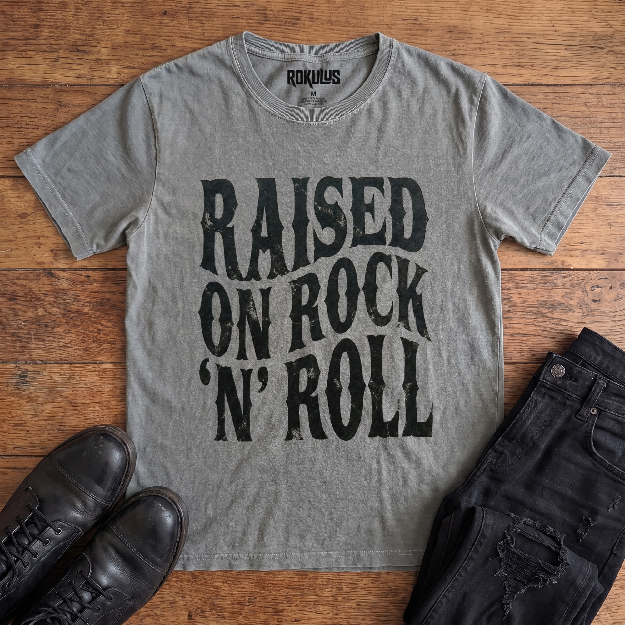 Washed grey Rokulus Raised On Rock 'N' Roll vintage graphic tee laid flat on wood with black boots and jeans, front print showing "Raised On Rock 'N' Roll" stacked in three lines of heavy wavy distressed black lettering
