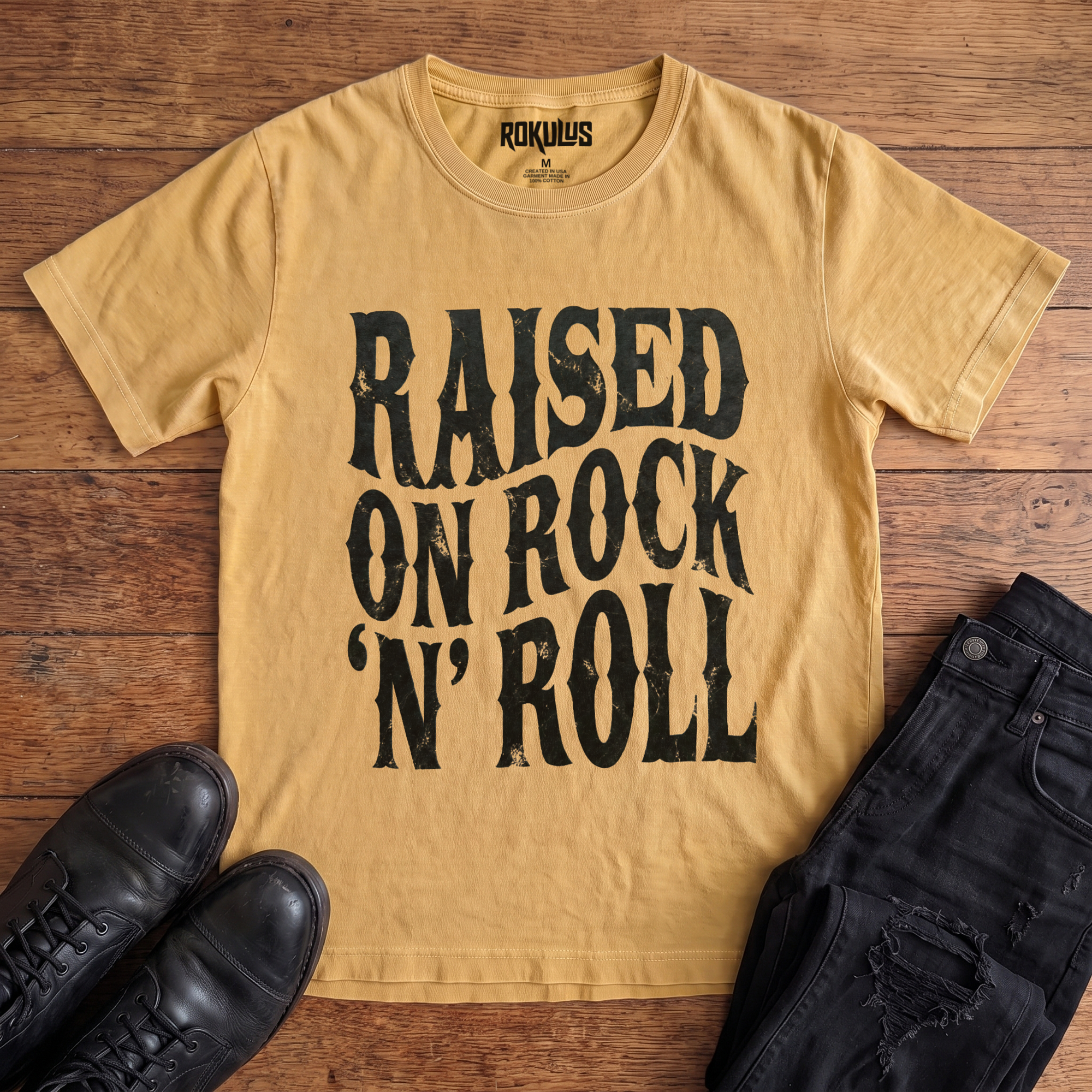 Washed mustard Rokulus Raised On Rock 'N' Roll vintage graphic tee laid flat on wood with black boots and jeans, front print showing "Raised On Rock 'N' Roll" stacked in three lines of heavy wavy distressed black lettering