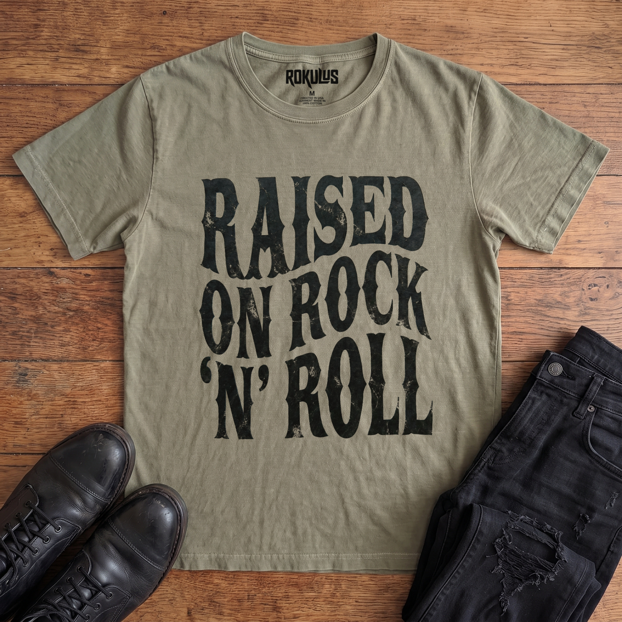 Washed olive Rokulus Raised On Rock 'N' Roll vintage graphic tee laid flat on wood with black boots and jeans, front print showing "Raised On Rock 'N' Roll" stacked in three lines of heavy wavy distressed black lettering