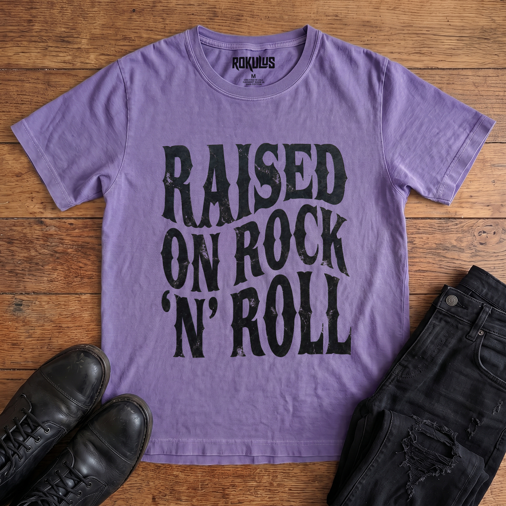 Washed purple Rokulus Raised On Rock 'N' Roll vintage graphic tee laid flat on wood with black boots and jeans, front print showing "Raised On Rock 'N' Roll" stacked in three lines of heavy wavy distressed black lettering