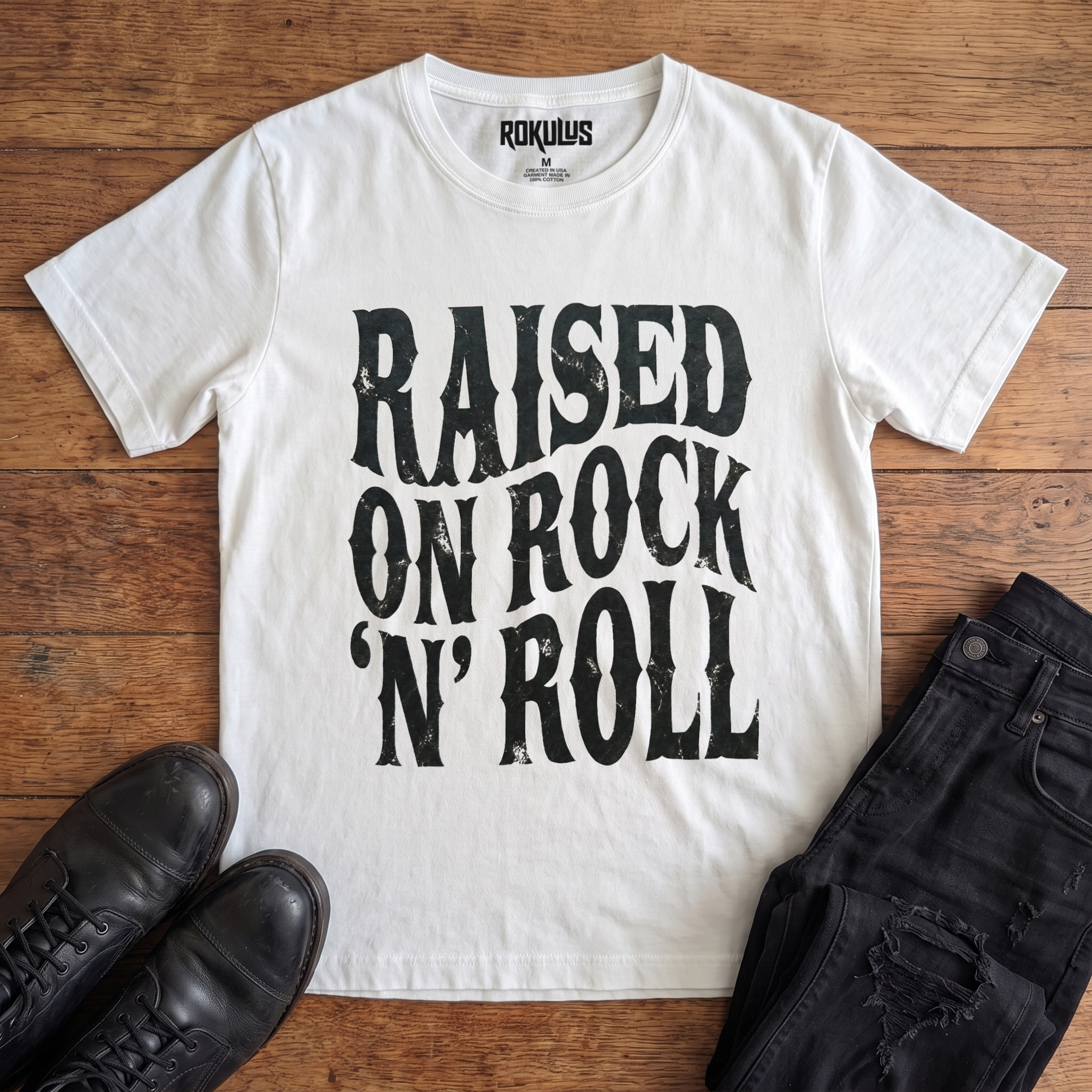 White Rokulus Raised On Rock 'N' Roll vintage graphic tee laid flat on wood with black boots and jeans, front print showing "Raised On Rock 'N' Roll" stacked in three lines of heavy wavy distressed black lettering