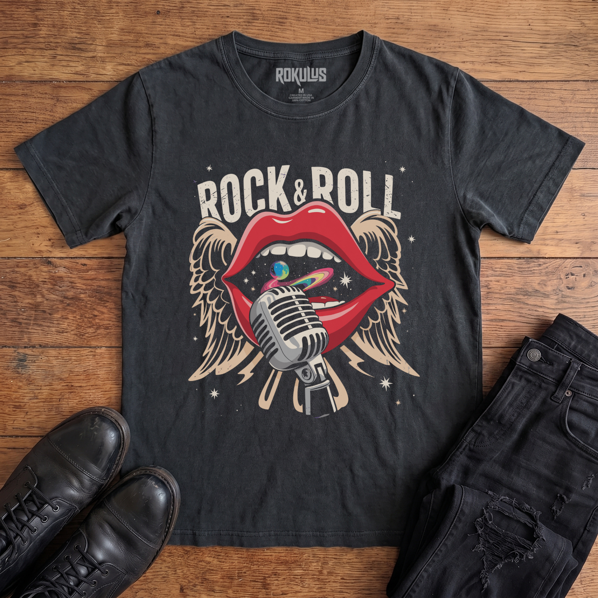 Washed black Rokulus Rock & Roll Lips vintage graphic tee laid flat on wood with black boots and jeans, front print showing bright red lips around a chrome microphone with cosmic swirl and cream angel wings under arched "Rock & Roll" lettering