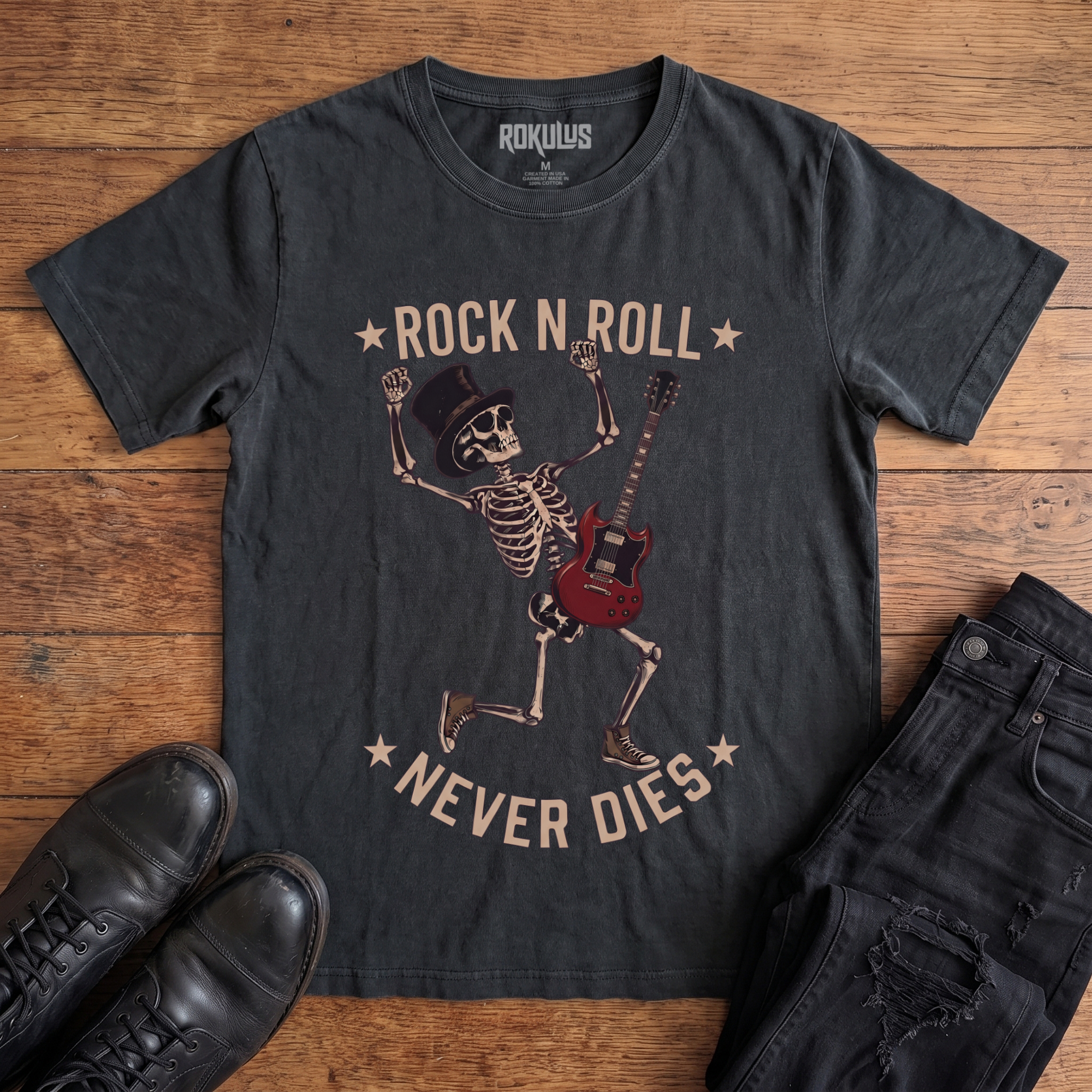 Washed black Rokulus Rock N Roll Never Dies vintage graphic tee laid flat on wood with black boots and jeans, front print showing a dancing top-hat skeleton with a red electric guitar and fists raised under arched "Rock N Roll Never Dies" lettering
